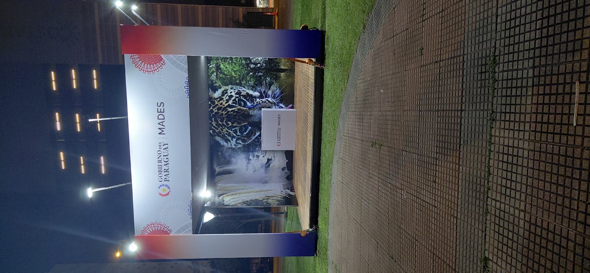 The image depicts an outdoor scene at night, likely at an event or exhibition in Encarnación, Paraguay. The focal point is a large display banner featuring a jaguar and a waterfall, with accompanying text. The banner is illuminated, creating a striking visual against the dark background.
To the left of the banner, a series of bright lights are visible, suggesting artificial lighting for the event or a nearby structure. The banner itself has a gradient of red, white, and blue at the top and bottom. Prominently displayed on the left side of the banner is the text "GOBIERNO DEL PARAGUAY MADES" in bold lettering. Below "GOBIERNO DEL PARAGUAY" is the national emblem of Paraguay, a star within a circular wreath. A smaller white box with the same text and emblem is placed on the display in front of the jaguar image.
To the right of the banner, a neatly paved walkway made of square tiles extends into the background. Adjacent to the walkway is a well-maintained grassy area. The overall impression is of a well-organized outdoor setting, possibly a public space or a venue for an official event. The time of day is clearly night due to the artificial lighting and the dark sky. No specific weather conditions are evident, but the grass appears green and healthy, suggesting it is not actively raining. The scene is calm and illuminated, setting a professional and visually appealing atmosphere.
