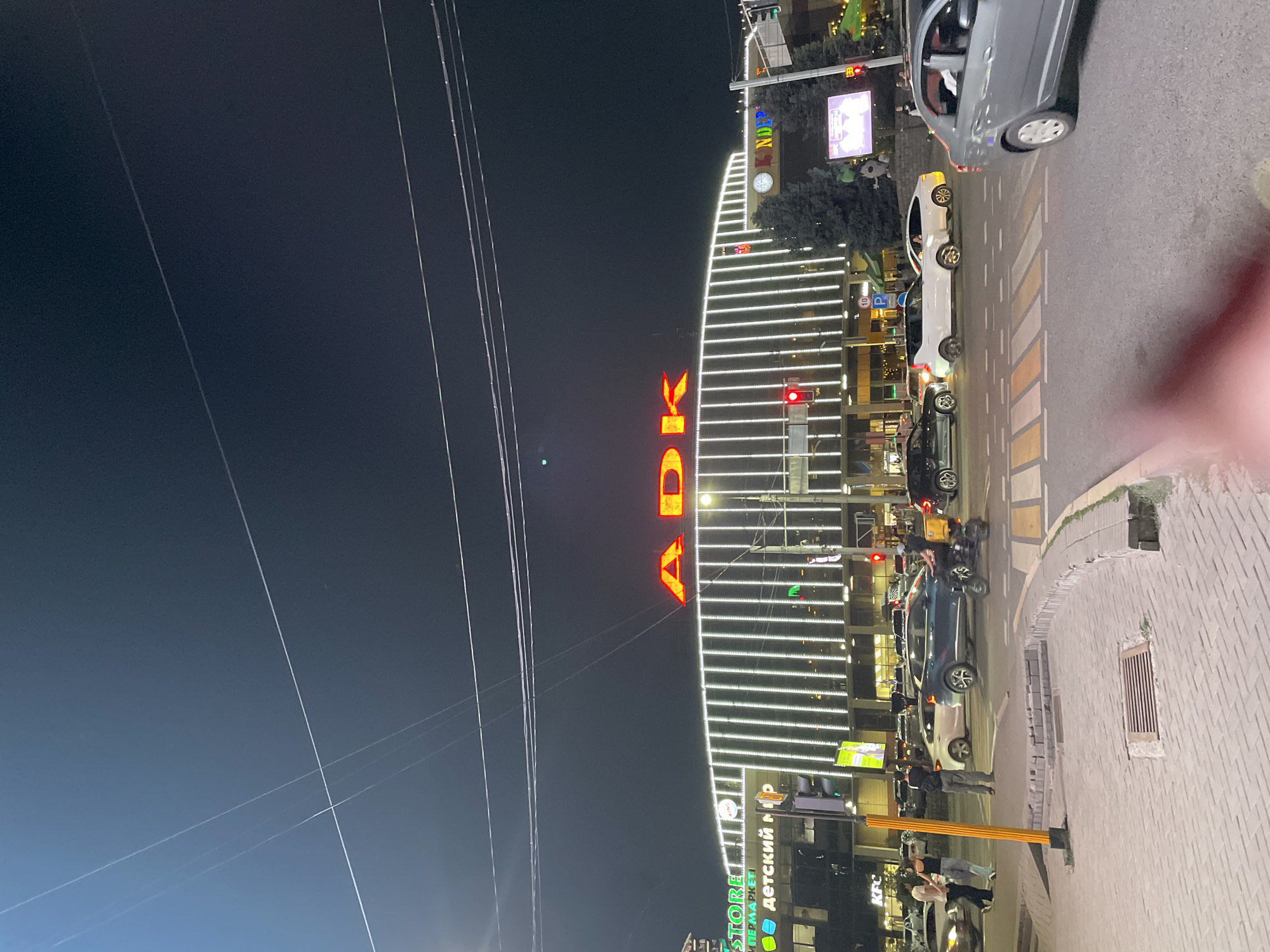 The image shows a nighttime view of the ADK shopping center, a prominent landmark located in Almaty, Kazakhstan. The building is illuminated with bright white lights along its facade, and the large, red "ADK" lettering is clearly visible against the dark sky. The street in front of the shopping center is busy with traffic, including cars and what appears to be a delivery vehicle with a yellow box. Pedestrians are visible on the sidewalks and crossing the street. Signs for "STORE," "KINDER," and "ДЕТСКИЙ МИР" are also visible on the building, indicating various shops within. The presence of traffic lights and streetlights suggests a typical urban intersection. The overall scene depicts a bustling commercial area at night.