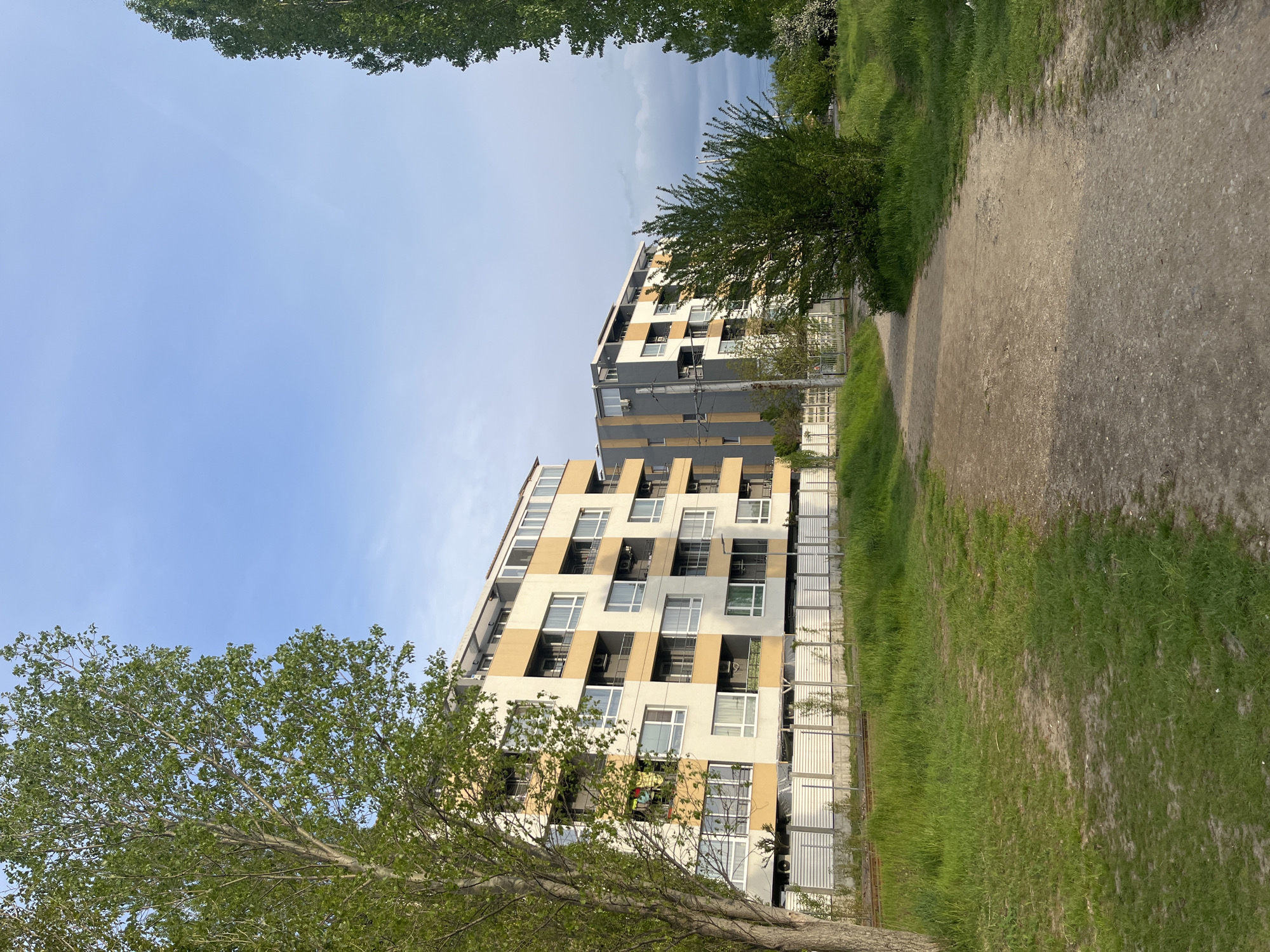 The image displays an outdoor urban landscape in Sofia, Bulgaria, featuring a combination of modern residential buildings and natural green spaces.
In the foreground, a light brown, unpaved dirt path winds from the bottom right towards the center of the frame. It is bordered on both sides by vibrant green grass, indicating a well-used but informal route.
Two multi-story apartment buildings stand prominently in the mid-ground. The building on the left exhibits a contemporary design with a facade featuring a mix of cream, beige, and white panels, interspersed with dark grey sections. It is adorned with numerous large windows and balconies. Further back and slightly to the right, a second, taller apartment complex is visible, characterized by a darker grey and black color scheme with contrasting yellow accents. Both structures appear modern and well-maintained.
Lush green trees are scattered throughout the scene. A large tree with abundant foliage occupies the lower left, its branches extending into the frame. Other trees line the path and are seen near the buildings, providing a natural buffer. Metal fences are visible along the edges of the property and beside the path, suggesting boundaries or security.
The sky above is mostly clear and bright blue, suggesting good weather, although a patch of darker, possibly overcast or distant stormy clouds is noticeable in the upper right. The lighting indicates it is daytime, likely morning or afternoon. There are no people or specific activities depicted. No visible text is present in the image.