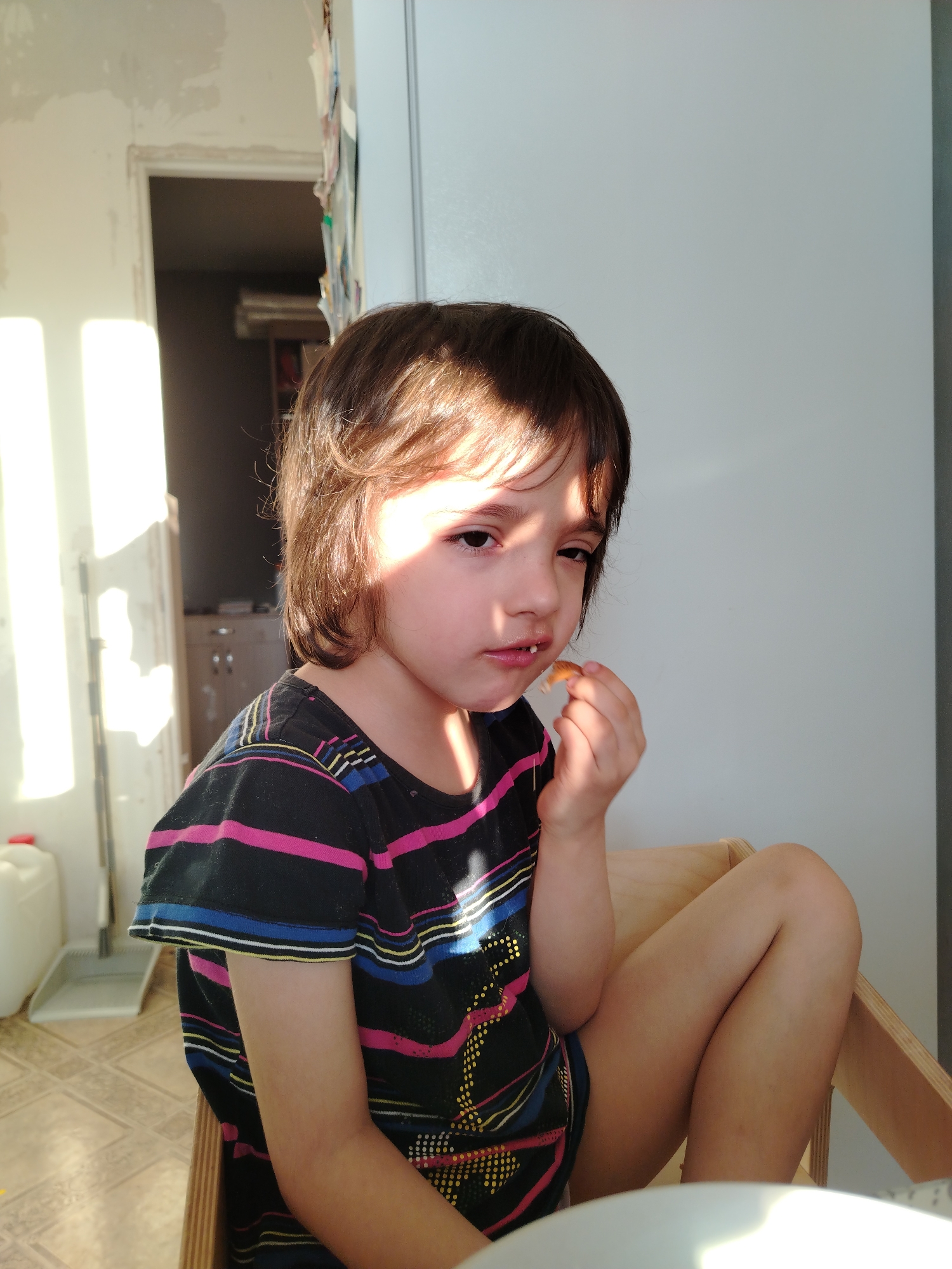 A young child with brown hair, wearing a dark short-sleeved shirt with pink, yellow and blue stripes, sits in a wooden chair and eats. The child's face is partially obscured by sunlight. The location appears to be indoors, possibly a kitchen or dining area, with a glimpse of another room visible through a doorway in the background. A white container and cleaning supplies are visible on the floor to the left. A white refrigerator or cabinet is on the right. There is no information to indicate that the location is in the city of Noviy, Russia.