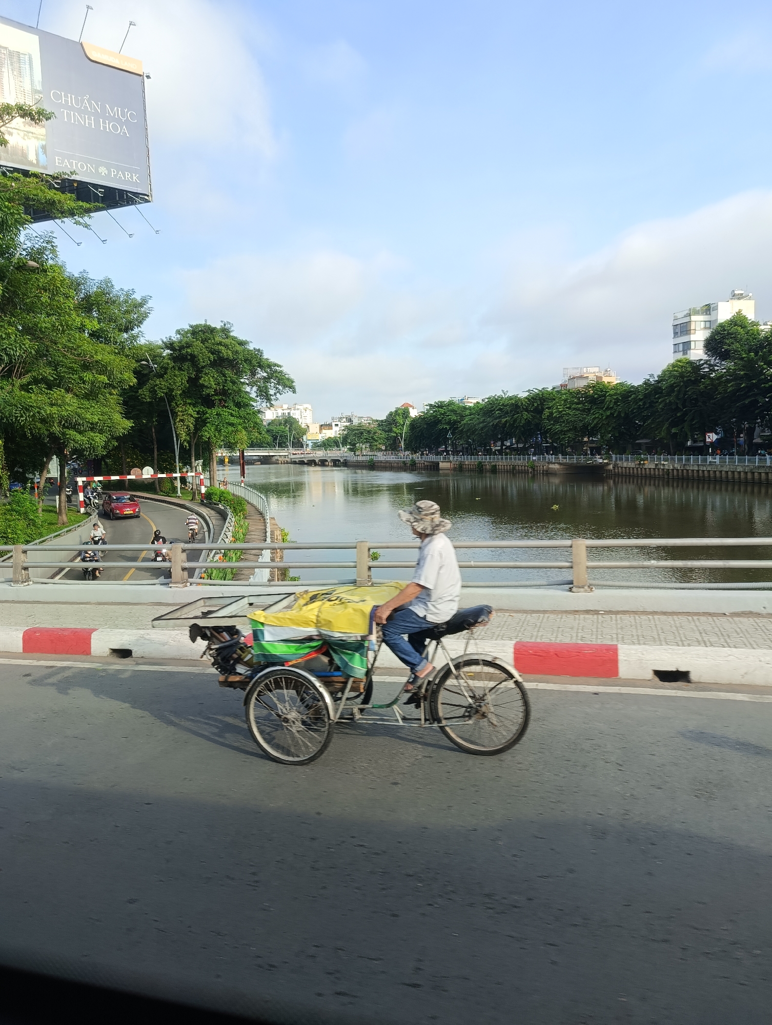 A man wearing a patterned hat and blue jeans is riding a three-wheeled bicycle, often referred to as a cyclos or pedicab, across a bridge over a river in Thủ Đức City, Vietnam. The cyclos is loaded with yellow and green tarps. The man is facing away from the camera. In the background, there is a tree-lined river with buildings and a bridge in the distance. The sky is partly cloudy. The scene appears to be an urban environment with traffic and pedestrians visible on the road and sidewalk. The overall atmosphere suggests a typical day in the city.