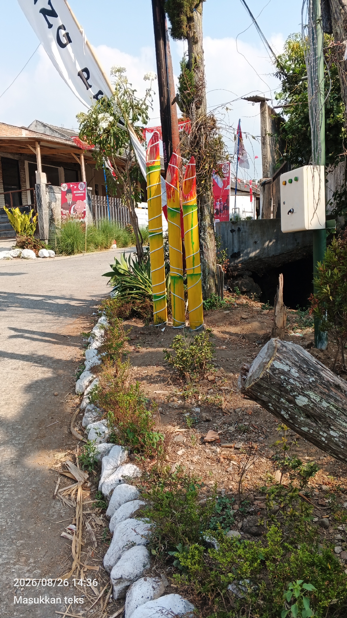 A street scene in Jeru, Indonesia, features three large, yellow bamboo poles painted to resemble pencils, adorned with green and red wrapping. They are positioned vertically next to a utility pole. To the left, a white banner with black lettering, partially obscured, hangs across the street. Red and white flags are visible further down the road, along with posters depicting people. The ground is a mix of dirt and asphalt, with a low stone wall lined with plants bordering the roadway. A large, fallen tree trunk lies on the right side of the image. A white electrical box with green buttons is mounted on a pole to the right of the "pencils." The image is dated 2026/08/26 14:36.