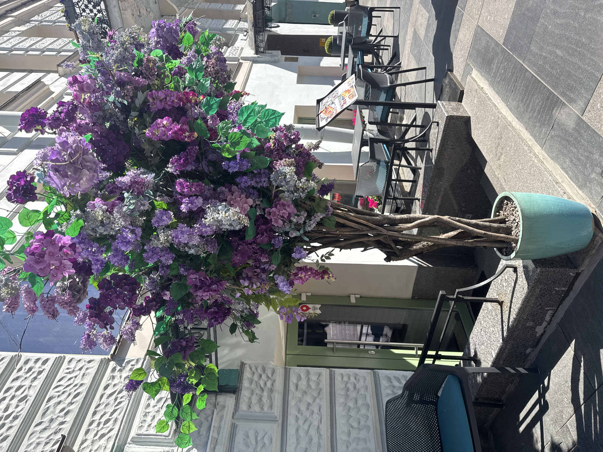 The image depicts an outdoor seating area of a cafe or restaurant, likely in Saint Petersburg, Russia. A large, decorative floral arrangement, predominantly featuring purple and lavender artificial flowers and green foliage, dominates the left side of the frame, partially obscuring the building facade. The building features ornate white stone detailing on the lower levels and a white upper story. A green door with a glass panel is visible, with a circular red and white sign above it.
The cafe seating consists of dark metal chairs with blue cushions, arranged around small tables. These are set on a patterned tiled floor, with shadows cast by the chairs and a nearby bench indicating a sunny day. The sun's angle suggests it is late morning or early afternoon. The ground and building materials appear consistent with an urban European setting.
There are no people clearly visible in the image, though reflections in the glass door suggest there might be activity inside. The overall impression is of a charming and inviting outdoor space, enhanced by the vibrant floral display. The weather appears to be fair and sunny.
Visible text includes a sign on an easel in front of the seating area, which appears to be a menu with images of food. A small red and white circular sign above the door has a pin-like graphic with an "i" in the center, commonly used to indicate information or a point of interest.