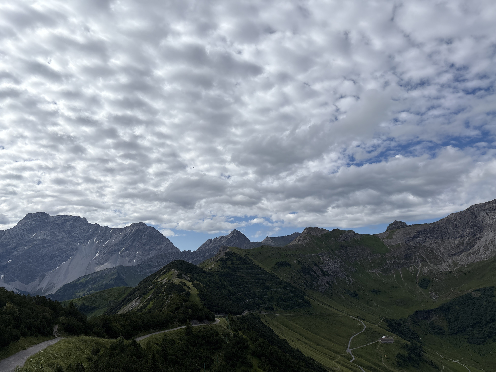 This is a landscape photograph taken from a high vantage point in Malbun, Liechtenstein, looking out over a vast mountain range under a cloudy sky. The foreground features a winding gravel road with sparse vegetation on either side. The middle ground consists of steep, green mountain slopes, some of which are terraced with rows of trees or vegetation. A few scattered buildings, possibly mountain huts or farms, are visible in the valleys. The background is dominated by imposing, rocky mountain peaks, some of which are partially covered in snow or scree. The sky is filled with a dense layer of cumulus clouds, with patches of blue sky visible in some areas. There are no people or animals visible in the image.