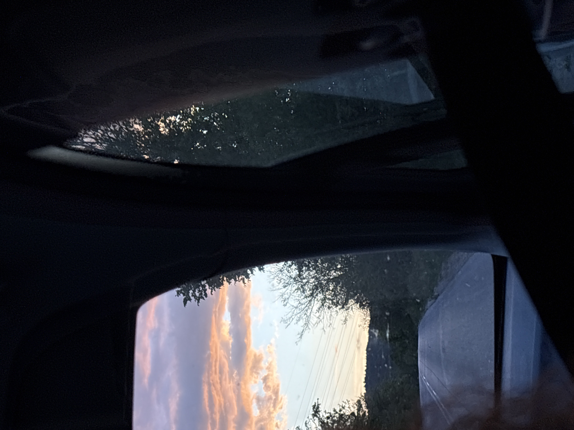 The image shows a view from inside a vehicle, likely a car, looking out through the sunroof and side window. The sunroof offers a view of trees, possibly silhouetted against a light source. The side window captures a scene of a pastel sky, with clouds tinged with orange or pink, likely at sunrise or sunset. Utility lines are visible in the side window. The surrounding environment outside the windows suggests an outdoor, potentially rural or suburban area. The interior of the car is dark, with the focus on the exterior scenes visible through the glass. It is not possible to determine the location as Bata, Bulgaria, from the image.