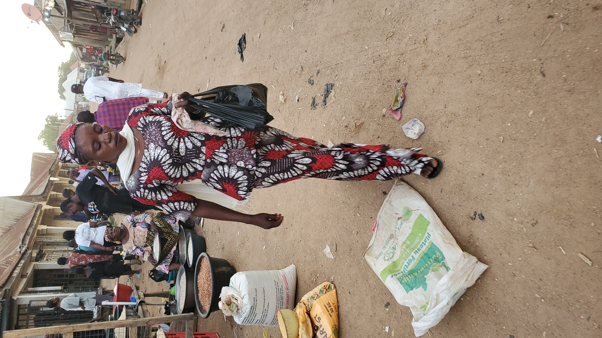 In Jalingo, Nigeria, a bustling marketplace is depicted with a strong focus on a woman in the foreground. She wears a vibrant, patterned outfit consisting of a long, flowing tunic with bold red, white, and black floral designs and a matching headwrap. A white scarf is draped around her neck, and she carries a black plastic bag in her right hand. Her gaze is directed downwards, and she appears to be walking away from the viewer.
Behind her, the marketplace is a hive of activity. Several other individuals are visible, some dressed in traditional attire, others in more casual clothing. Stalls are set up along a dusty street, displaying various goods. Notably, large bowls filled with grains, possibly beans or lentils, are prominent. Sacks of what appear to be agricultural products, including one with "NET WEIGHT 50 KG" printed on it, are scattered on the ground. In the background, a row of single-story buildings with corrugated metal roofs lines the street, suggesting a typical urban market environment. Motorcycles are parked along the side of the road, indicating common modes of transport in the area. The overall impression is one of a lively and active market day.