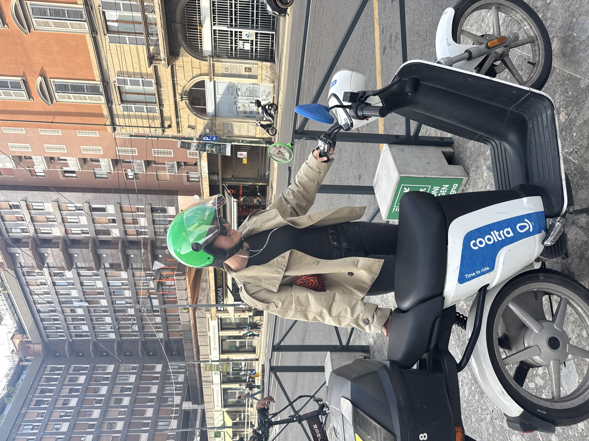 The image depicts a person on a Cooltra scooter, likely in an urban environment in Italy. The person is wearing a light brown jacket, black shirt, and a lime green helmet. The Cooltra scooter is primarily white with a blue "Cooltra" logo and the phrase "Time to ride."
The background shows buildings of varying heights and architectural styles, constructed from stone or brick. Multiple windows are visible in the buildings. Some street elements such as poles, signs, and other parked vehicles are present. The overall scene suggests the subject is on a street in a European city. The location is identified as Milan, Italy based on the signs in the background.