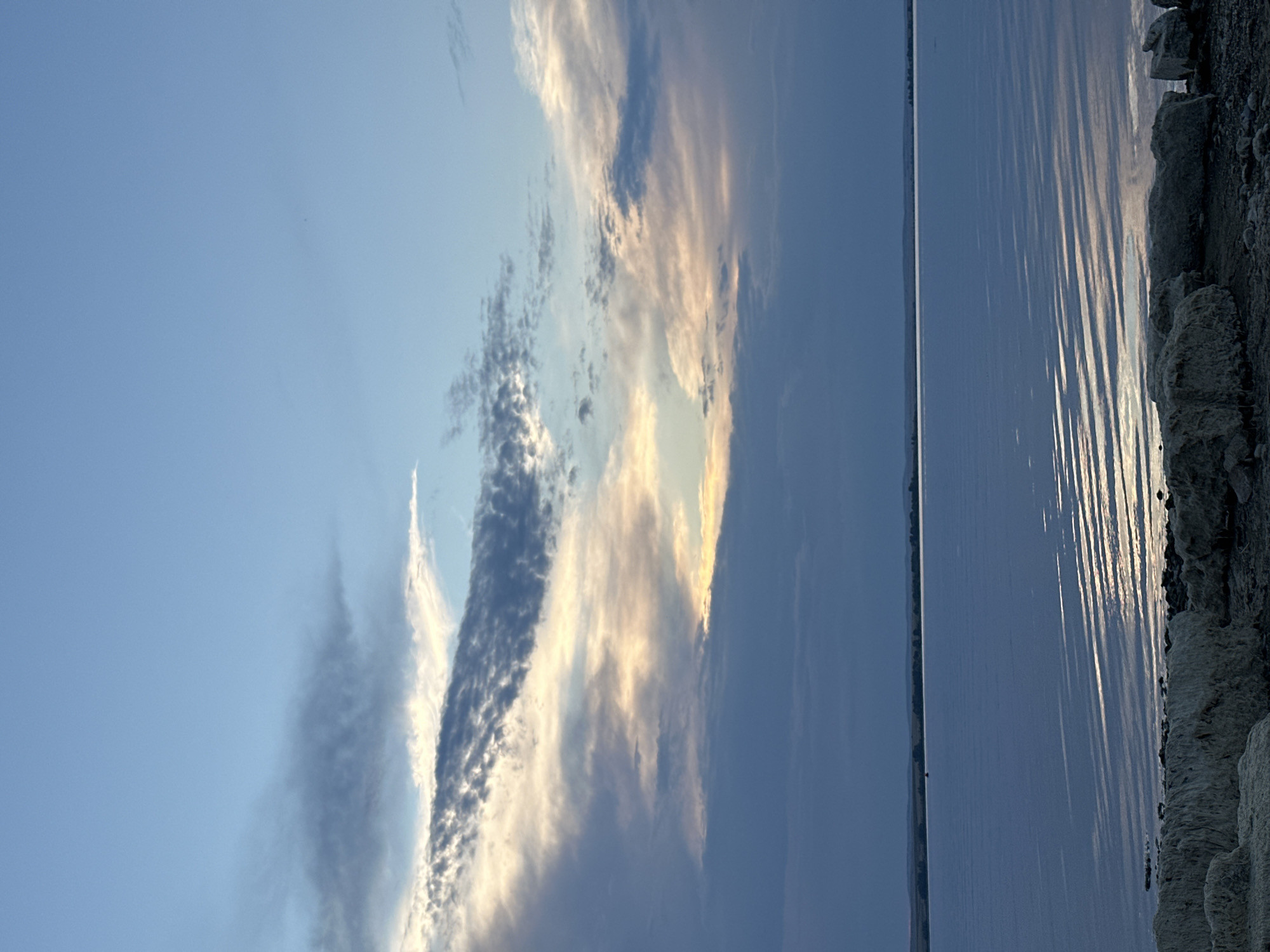 This image shows a body of water at dusk, with a rocky shoreline in the foreground and a silhouetted landscape in the distance. The sky is a soft blue with scattered clouds, some of which are lit by the setting sun. The water is calm, with ripples reflecting the light. The shoreline is made up of large, light-colored rocks. The scene appears to be a natural setting, possibly a lake or a large river. The location is not specified in the image, and therefore it cannot be identified as Energetik, Russia, or any other specific city.