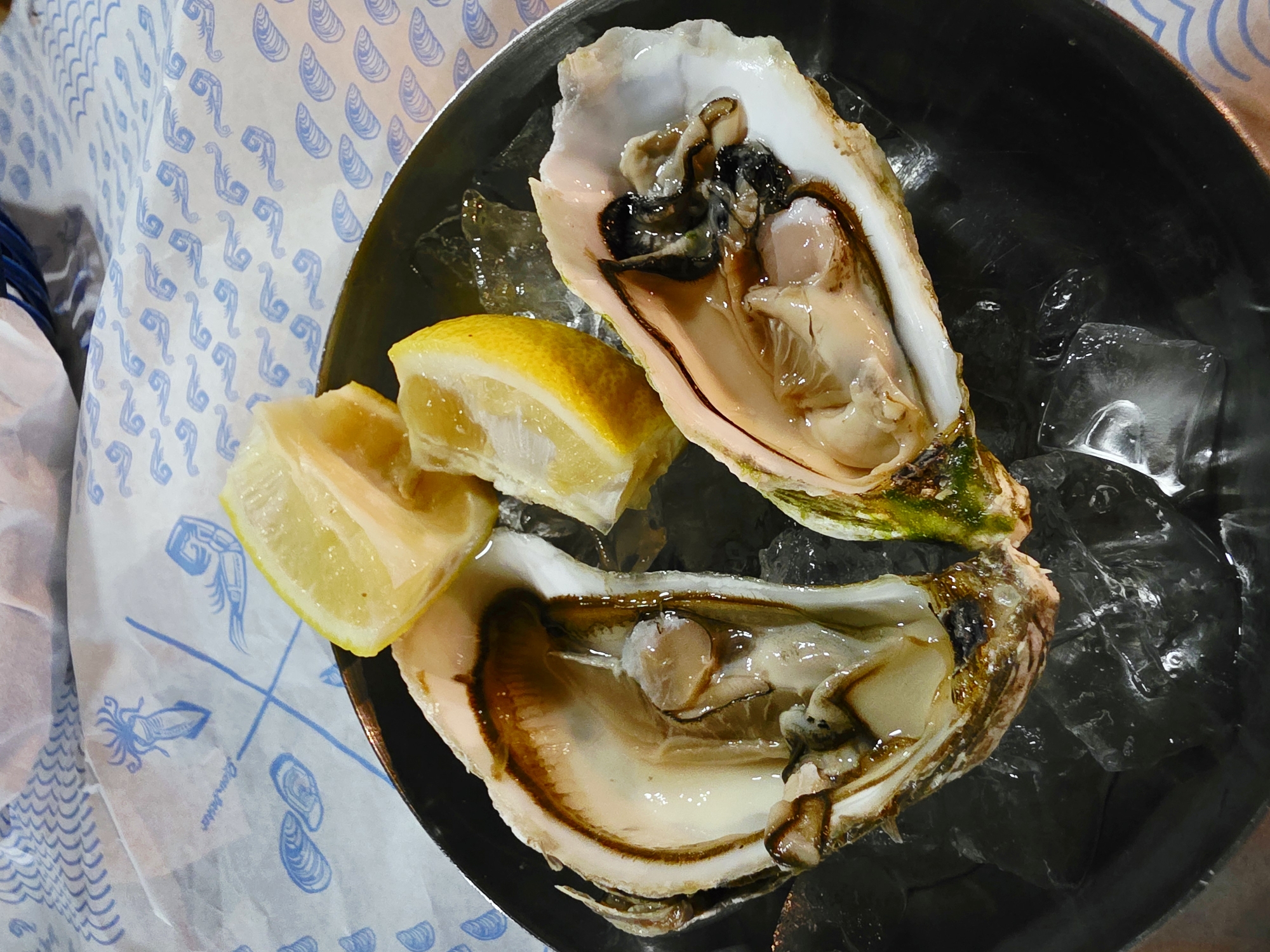 Two opened oysters are presented on a bed of ice within a dark-colored serving dish. The oysters are garnished with three lemon wedges. The dish is placed on a white paper liner adorned with a blue pattern of stylized sea creatures and shells. The scene appears to be an outdoor setting, possibly at a restaurant in Larnaca, Cyprus, given the context. There are no people visible in the image.