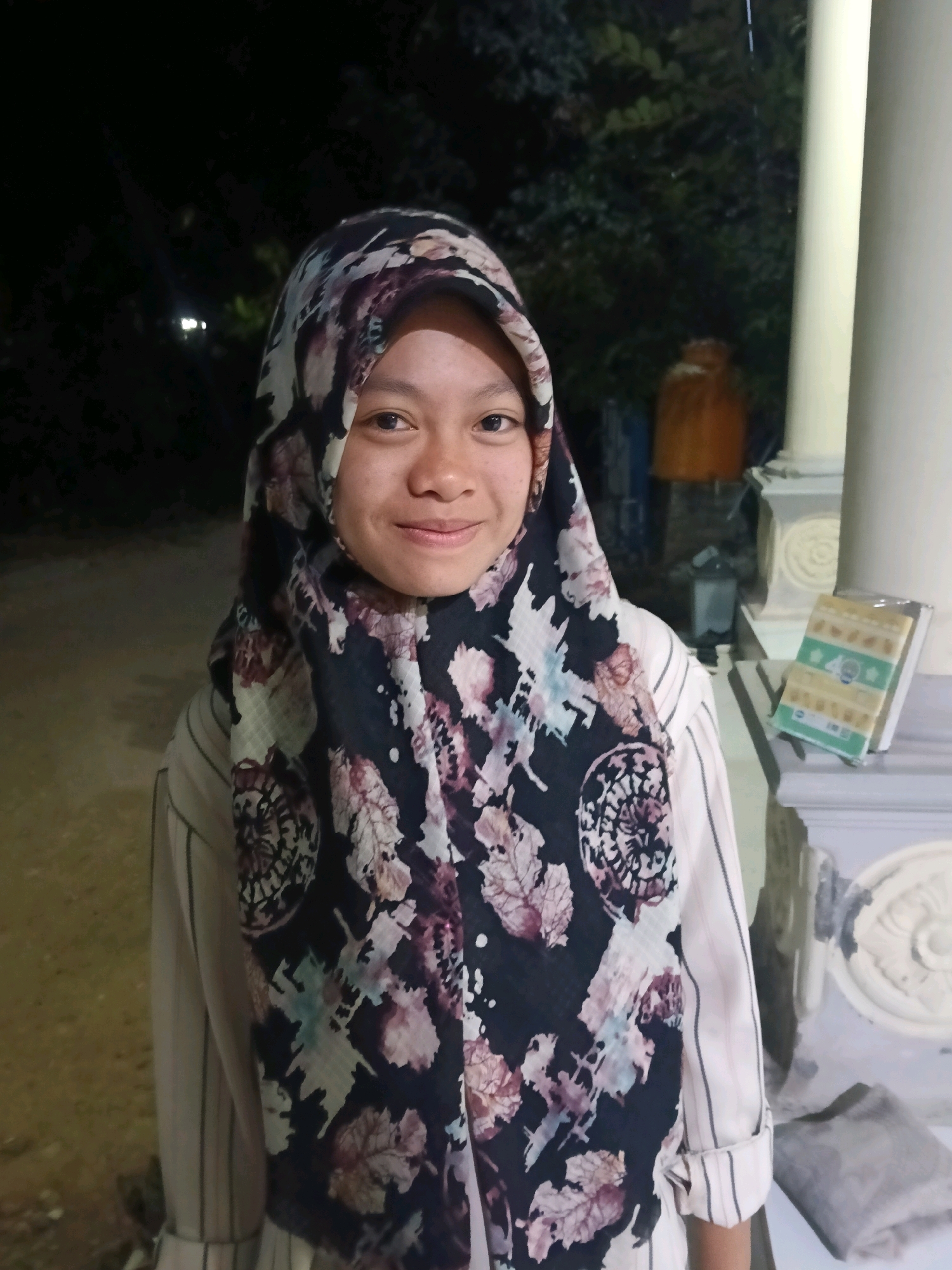 A young woman wearing a floral hijab and a striped shirt stands outdoors at night. She is looking at the camera and smiling. Behind her is a dark background with trees and some structures. To her right is a white pillar and a shelf with a book or religious text. The location is identified as Bulangan Barat, Indonesia. The image has a slight grainy quality, suggesting it was taken in low light conditions.