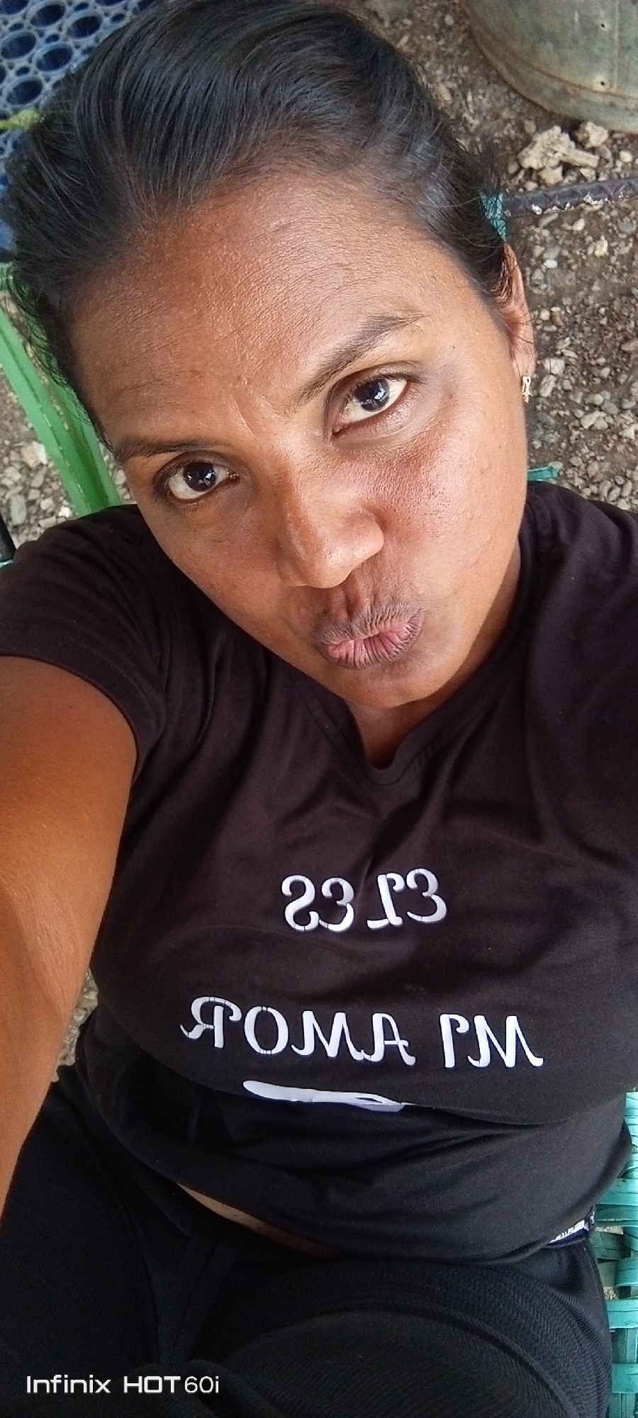 A woman is centered, angled slightly, from the chest up. She purses her lips in a kiss. The woman has dark hair pulled back, dark eyes, and dark skin. She wears a black short-sleeved t-shirt and black bottoms. The phrase “AMOR PM” is printed in white letters on the shirt, with another phrase above it that is backwards. She appears to be sitting on a green and blue plastic chair. The background shows what appears to be some type of debris or dirt and a partially visible bucket. The bottom of the image reads “Infinix HOT 60i”. The location of Altagracia, Venezuela, is unknown from the image.