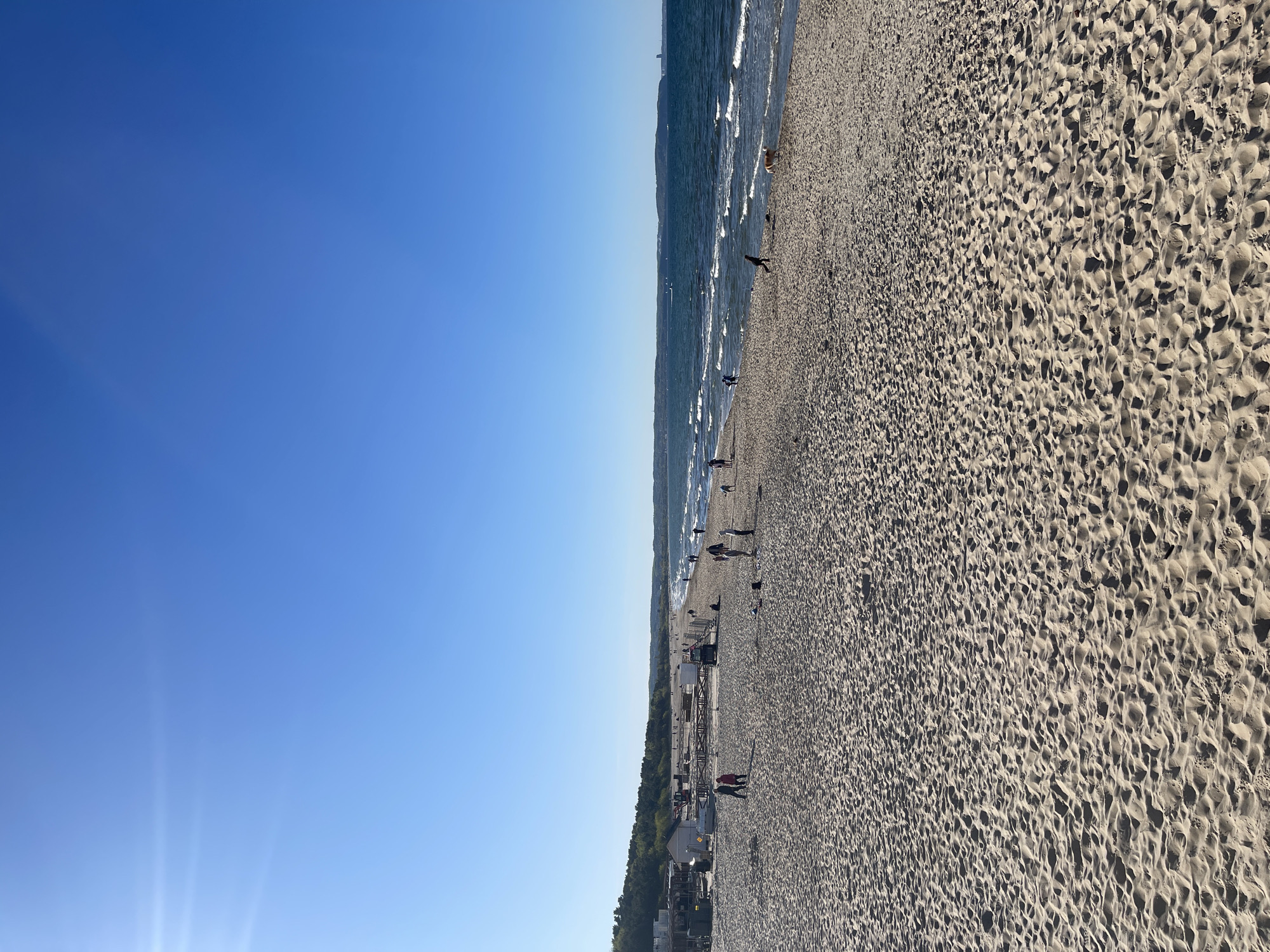 The image shows a beach in Gdańsk, Poland, on a clear, sunny day. The sky is blue and cloudless. The sandy beach stretches from the lower right corner to the upper right corner, with visible footprints in the sand. The Baltic Sea is along the upper center, with gentle waves lapping the shore. People are seen walking and standing along the shoreline, scattered throughout the beach. A line of trees and low buildings are visible in the middle of the image, separating the beach from the inland area.