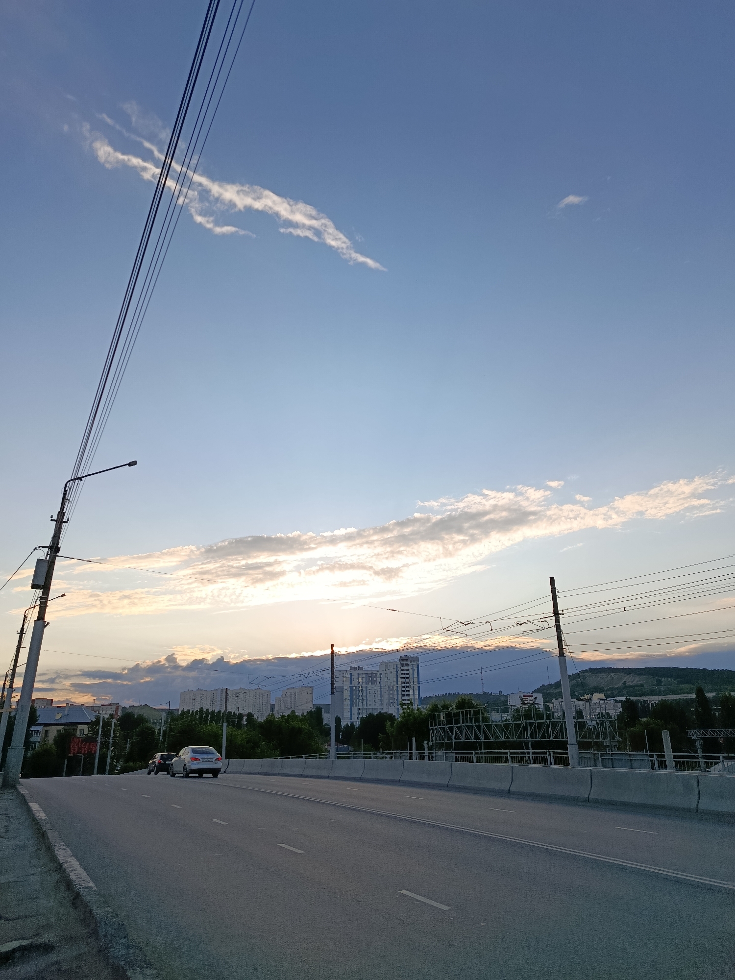 The image depicts a view of a road leading into a city, with a sunset casting a warm glow on the horizon. The road is paved and marked with white dashed lines. On the right side of the road, a concrete barrier separates it from a lower level, possibly another road or railway. In the distance, several multi-story buildings and some trees can be seen. Overhead, power lines stretch across the frame, supported by utility poles. The sky is a clear blue with some wispy clouds. Two cars are visible on the road, one white and one dark-colored, both moving away from the viewer. The overall scene suggests an urban environment during the late afternoon or early evening.
This image shows the Komsomolsky Bridge in Saratov, Russia, at sunset. The bridge is a road bridge spanning the Volga River, and the view is of the approach to the city. The buildings in the background are typical of Soviet-era apartment blocks, and the power lines are part of the city's infrastructure. The sunset creates a dramatic backdrop, with the warm light illuminating the clouds and casting long shadows.