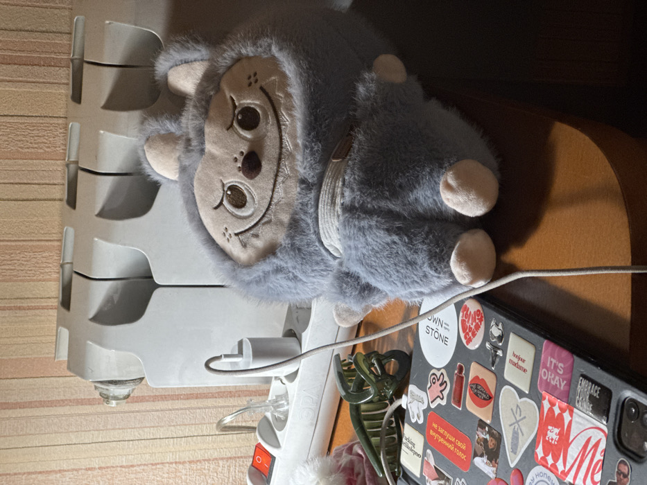 A gray plush toy with an embroidered face sits on top of a white power strip and a dark-colored laptop covered in stickers. The laptop rests on a wooden surface. The stickers display various designs and phrases like "OWN = STONE," "IT'S OKAY," "EMBRACE YOUR CRINGE," and a Russian phrase. A green hair clip is also visible near the laptop. A white radiator is in the background, adjacent to a patterned wall. A white cable extends from the power strip towards the laptop.