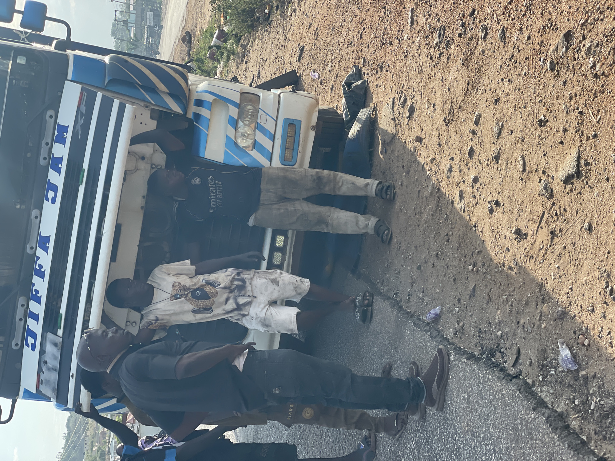 The image depicts a group of men standing around the front of a large truck with "JIFFA" printed vertically on the side. The truck is white with blue stripes, and the front appears damaged. The men are dressed in casual clothing, including t-shirts, shorts, and trousers. One man wears a black shirt with "Emirates | Fly Better" written on it. The location appears to be alongside a road, with a dirt shoulder and some vegetation. A glimpse of buildings and structures in the background suggests an urban setting. The image was taken in Lokoja, Nigeria.