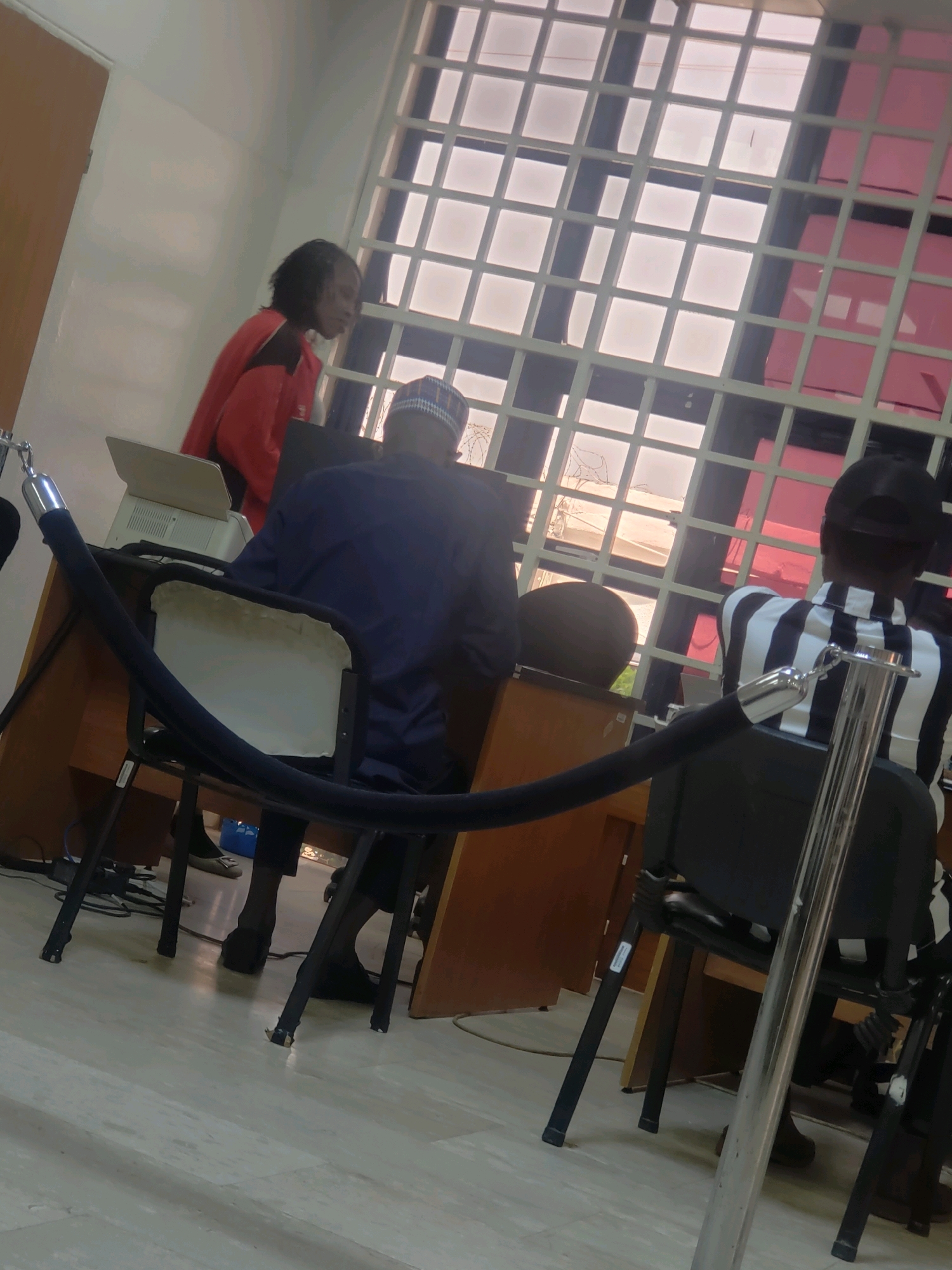 In Kakuri Gwari, Nigeria, an indoor scene depicts a service counter area. A woman in a red jacket stands to the left, interacting with a person behind a desk. A man wearing a blue shirt and cap is seated at the desk, and another individual in a black and white striped shirt is seated to the right, facing away from the camera. The environment includes a desk with a printer, chairs, and stanchions with a velvet rope, suggesting a queue management system. A large window with a grid pattern is visible in the background. The flooring is tiled, and wooden doors are present on the left side of the frame.