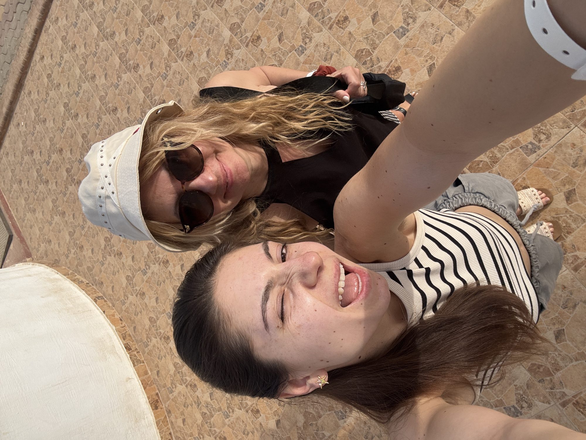 Two women are posing for a selfie in an outdoor location. One woman is wearing a white bucket hat and sunglasses, and a black shirt. The other woman is wearing a black and white striped top. They are standing on a light brown tile surface with mosaic-like patterns. A partially visible white ledge is in the lower left corner. The city is unknown.