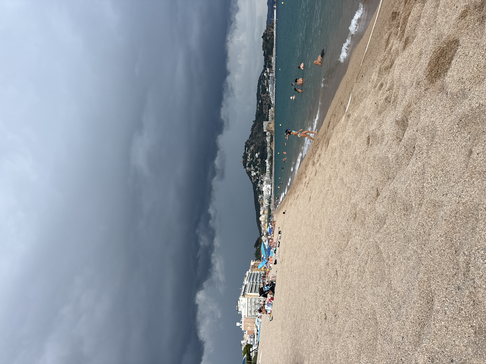 This image depicts a wide, sandy beach in Blanes, Spain, with the Mediterranean Sea stretching out to the horizon. The sky is dominated by heavy, dark storm clouds, casting a somber mood over the scene, though the water itself appears calm and blue.
On the beach, numerous people are visible, some sunbathing on towels or under umbrellas, while others are engaged in water activities. A group of individuals are swimming or wading in the sea. The shoreline is a mix of sand and small pebbles.
In the background, a picturesque coastal town rises up a hill, characterized by white buildings and a church or castle-like structure at the summit. The architecture suggests a Mediterranean setting. The sea is dotted with a few small boats. The overall impression is of a beach resort town preparing for or experiencing the onset of inclement weather.