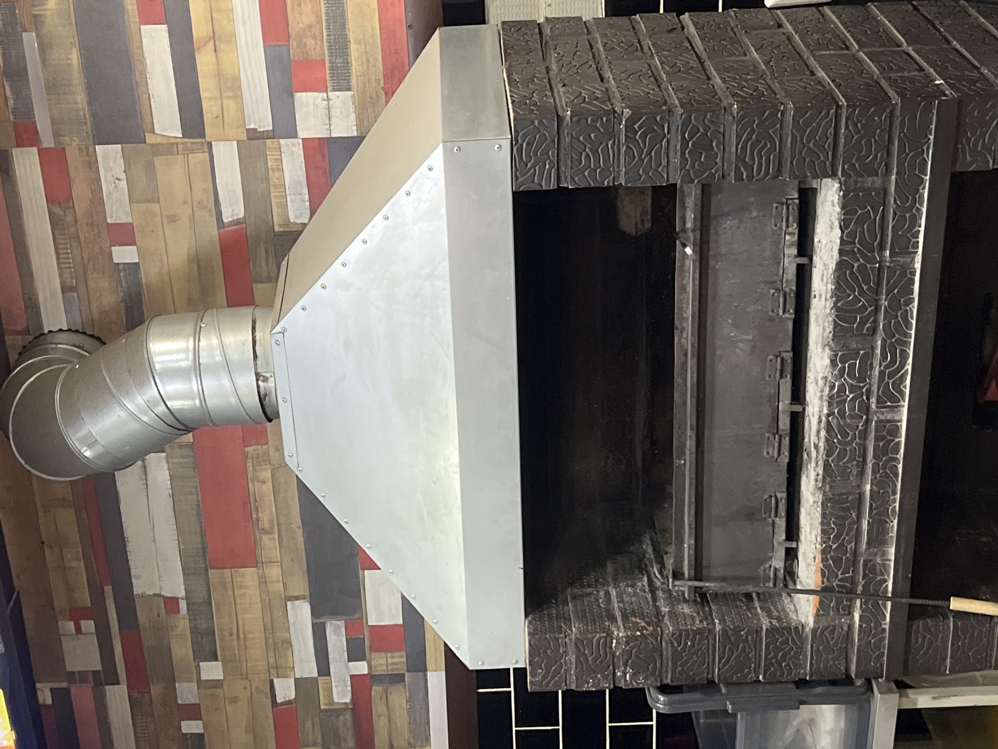 The image displays an indoor scene, likely from a commercial kitchen or restaurant in Chaikovsky, Russia, showcasing a cooking station with its associated exhaust system.
A large, dark-colored commercial oven or grill occupies the right side of the frame. Its facade is constructed from dark grey or black textured material, possibly decorative bricks or cast iron, adorned with an intricate, organic, relief pattern. An open section of the oven reveals a dark interior, within which a hinged metal door or baffle is visible. A long, thin metal poker with a light-colored wooden handle is positioned near the bottom right edge of the oven opening, suggesting preparation for cooking.
Above the oven, a substantial, industrial-style exhaust hood system is installed. It is made of light-colored, possibly galvanized, metal panels, fastened together with visible rivets, forming a broad, angled canopy. Extending from the left side of this hood is a corrugated, silver-colored metal flue pipe, which curves downwards and then horizontally out of view.
The background features a wall covered with rectangular panels resembling distressed wood planks. These planks are arranged in a varied pattern and come in an assortment of colors, including natural wood tones, white, red, and dark gray or black, creating a rustic or patchwork aesthetic. The scene is brightly lit, indicative of an indoor setting. No people or visible text are present in the image.