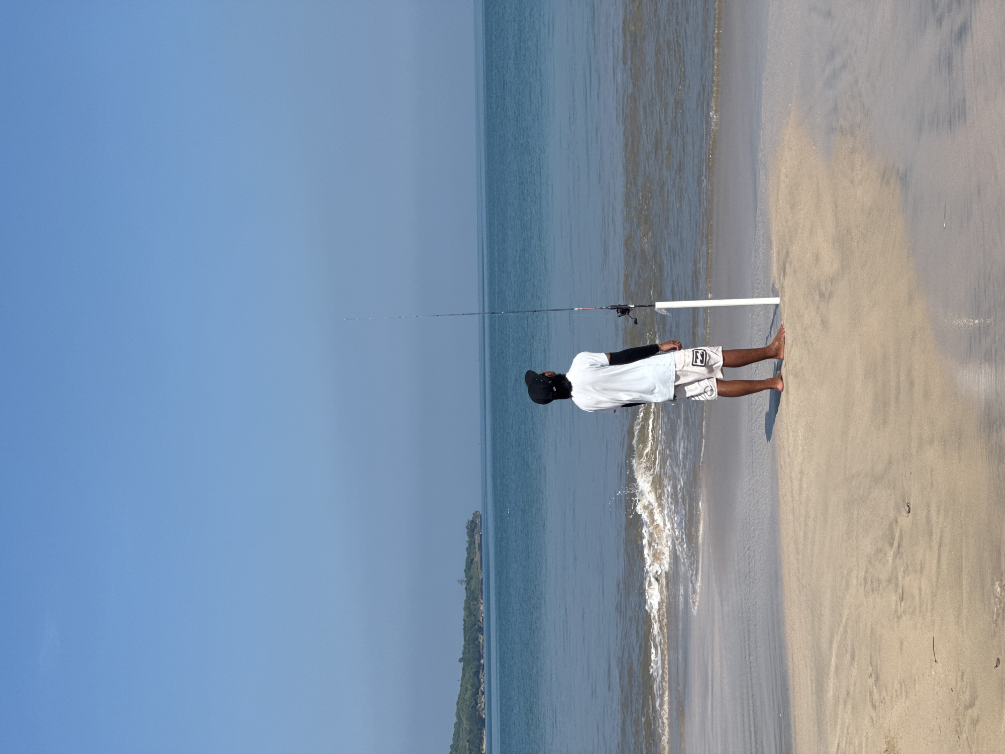 A man stands barefoot on a tan sand beach with a fishing pole extended over the water. He wears a white t-shirt, black cap, and patterned shorts. The water is light brown near the shore, transitioning to a dark blue further out. The sky is a clear, light blue. A line of trees runs along the coastline in the distance. This scene is likely in Kuta Selatan, Indonesia.