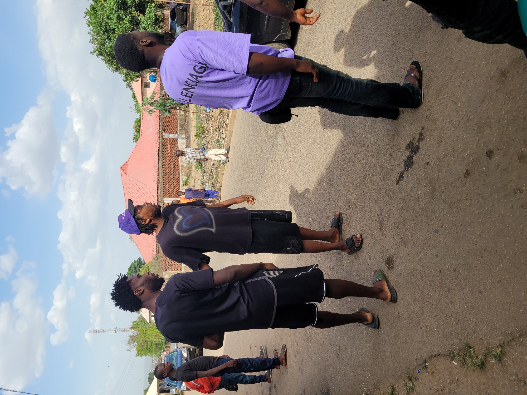 The image captures an outdoor scene featuring several young men standing on a paved street or pathway, likely in Jos, Nigeria. The photo is oriented ninety degrees clockwise, showing the subjects positioned sideways.
In the foreground, from left to right in the correctly oriented image:
A man in a dark t-shirt and dark shorts with white trim stands with his back partly to the camera, wearing dark green flip-flops.
Next to him, another man in a black t-shirt, black shorts, and black flip-flops is partially obscured.
The central figure is a man wearing a black t-shirt with a light-colored design, black shorts, and dark flip-flops. He has a purple baseball cap on and is looking upwards.
Further right, a man in a purple t-shirt with the word "BALENCIAGA" visible on the back, dark jeans, and dark flip-flops faces away from the camera, looking upwards and to the left of the frame. A small black pouch or bag hangs from his hip.
In the background, behind the subjects, are several buildings with red roofs, surrounded by green trees and foliage. A communication tower is visible in the distant left. In the mid-ground, a man in a plaid shirt and light pants stands near a building. Another person in a red top and blue jeans is visible in the lower left portion of the image, facing away.
Cars are visible in the background and foreground, including a black car near the man in the purple shirt and a blue car further down the road. The sky is bright blue with scattered white clouds, indicating daytime. Shadows are cast on the ground, suggesting a sunny day. The men appear to be casually standing or observing something in the distance or above them, as several are looking upwards.