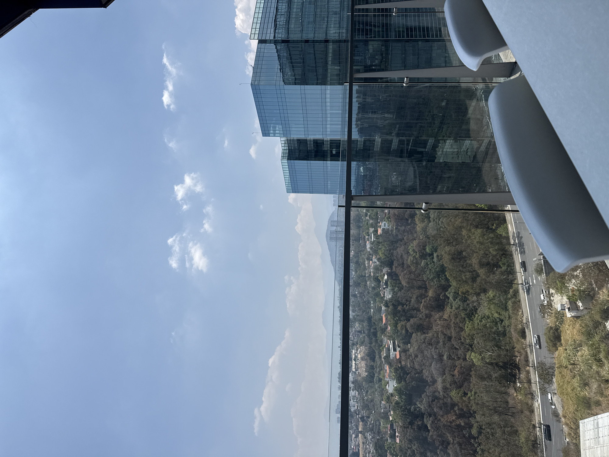 View from a high vantage point in Mexico City, Mexico. A modern glass skyscraper stands prominently. An expanse of cityscape and green space stretches out below, with mountains visible in the distance. Traffic moves along a highway. The sky is blue with scattered clouds. Modern white furniture is in the foreground.