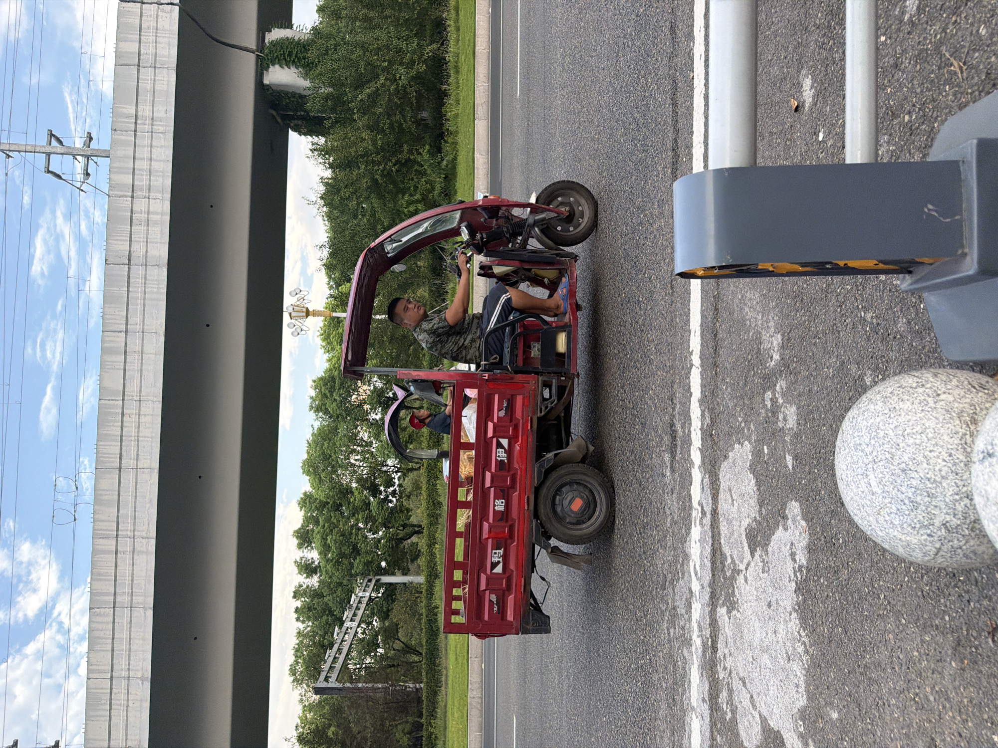A red, three-wheeled motor vehicle with a cargo bed is traveling on an asphalt road in the city of Jiangjin, China. A man wearing a camouflage t-shirt and shorts is driving the vehicle. He is wearing a red helmet. There is another person in the passenger seat of the vehicle, wearing a red helmet and a dark shirt. The cargo bed contains some yellow hay-like material and some plastic-wrapped items. The vehicle is under a large concrete overpass. Trees and power lines are visible in the background. In the foreground, there are decorative stone spheres and a grey traffic barrier. The sky is cloudy.
