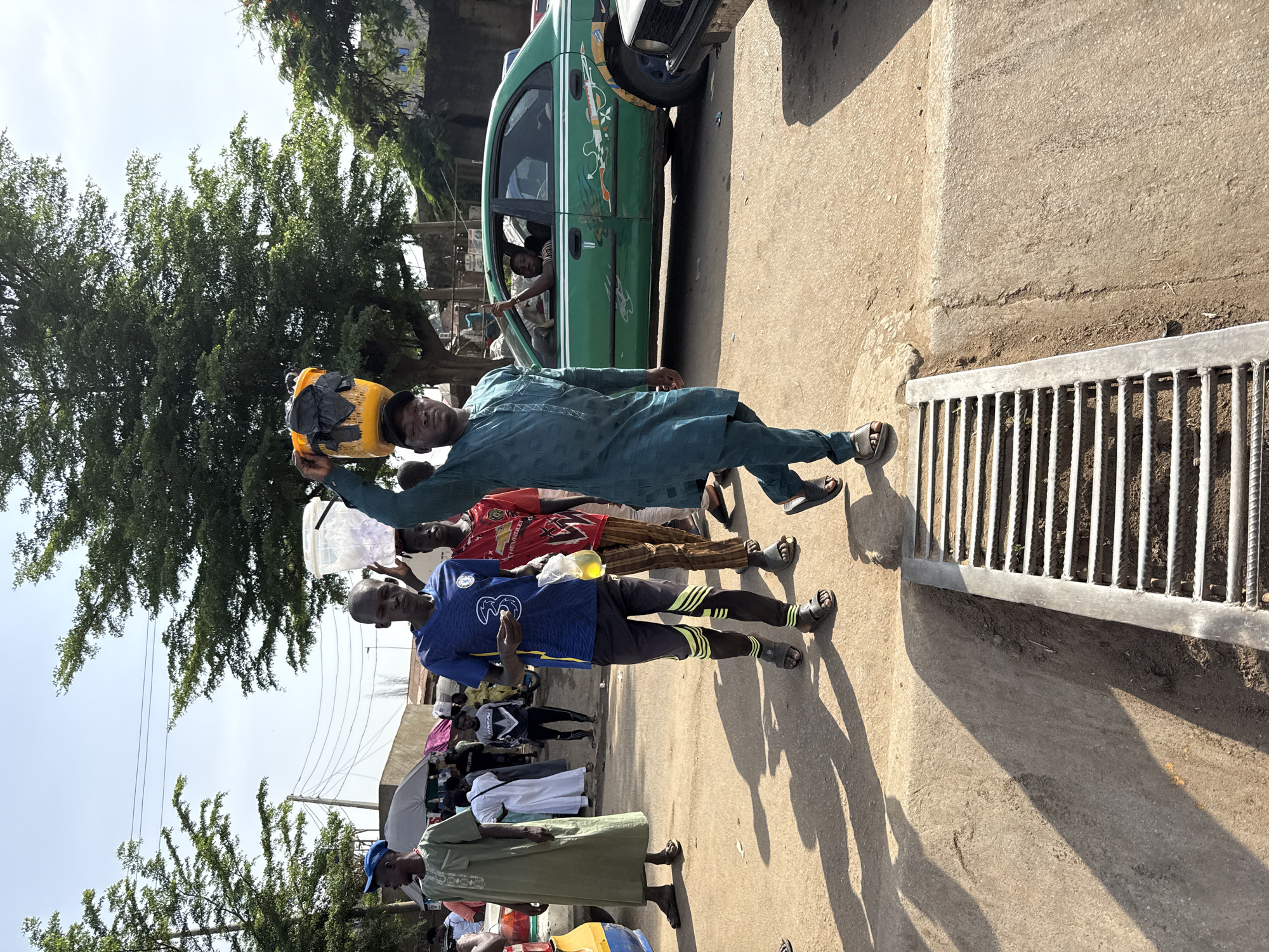 This image depicts a vibrant street scene, likely in Abuja, Nigeria, under bright daylight with strong shadows indicating a sunny sky. The central figure is a man dressed in a teal/green agbada (a traditional West African flowing garment) and a black cap. He is balancing a yellow jerrycan, possibly filled with water or another commodity, on his head with one hand, while his other arm is extended forward.
Behind him stands another man wearing a blue Chelsea FC football jersey, clearly displaying the number "3" and the "Chevrolet" logo, along with dark trousers featuring neon yellow stripes. He is holding a clear plastic bag containing a yellow liquid, which could be juice or a local food item, seemingly engaging in a transaction or interaction with the man in the agbada.
Further into the background, several other individuals are present: a person in a red patterned shirt, another in a long white garment, and one in a long green dress, all contributing to the busy atmosphere. To the left, a distinctive green car, possibly a taxi, with decorative white markings on its side, is visible, and a person's arm is extended from its open window.
On the right side of the paved ground, a metal drainage grate is visible. The background features lush green trees, blurred figures of people, some under umbrellas or makeshift shades, and hints of distant buildings, all under a clear sky with visible overhead power lines. The scene portrays typical daily life and street vending activity in an urban setting.