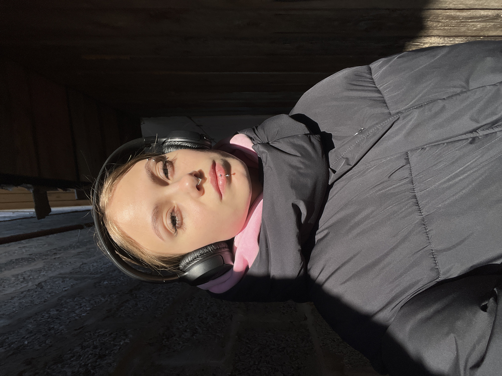 A fair-skinned person lies on a sunlit surface, possibly stone or textured ground, near a wooden structure, perhaps a building or fence. The person is wearing black headphones, a pink hooded garment, and a black padded jacket. The person has facial piercings including one in the septum and one above the right eyebrow. The surrounding environment appears to be shaded except for where the person is lying. There is no definitive evidence to confirm the location is Фоки, Russia.