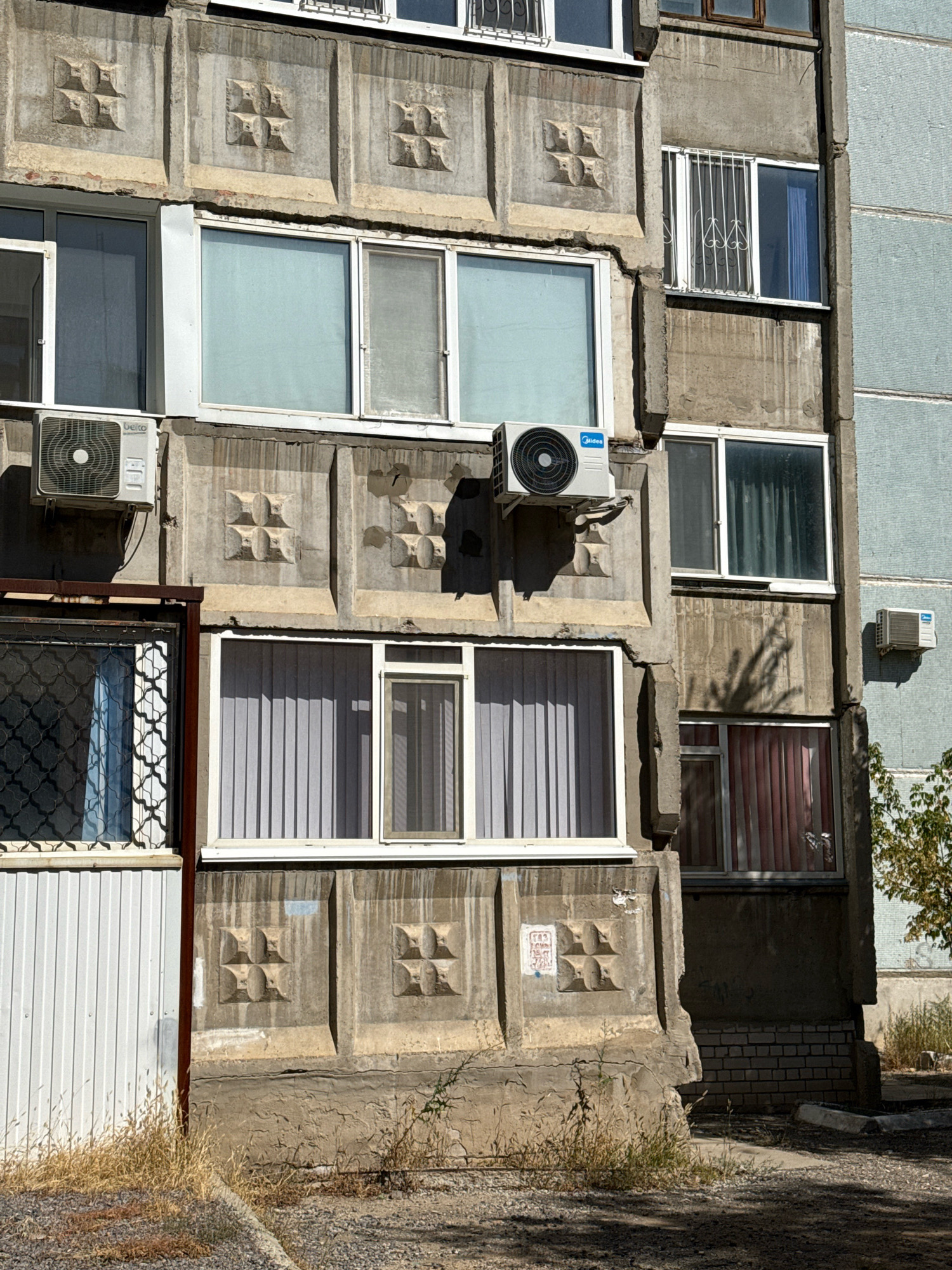 The image shows a close-up view of the exterior of a concrete apartment building in Aktobe, Kazakhstan. The building features a textured concrete facade with repeating square decorative elements, each containing a four-petal floral or geometric pattern. Several windows are visible, some with white frames and light blue or white curtains, and others with security bars. An outdoor air conditioning unit is mounted on the wall below one of the windows. The lower part of the building shows signs of wear and tear, with some cracks in the concrete and a section covered with corrugated metal siding. Dry grass and gravel are present in the foreground.