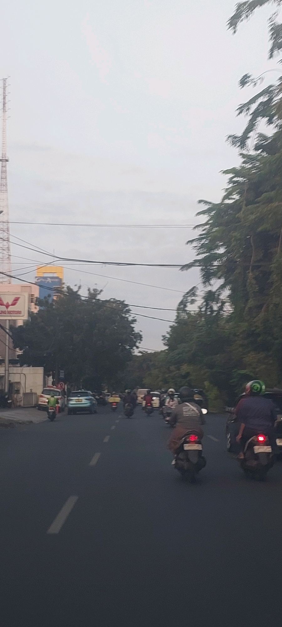 The image captures a street scene in Surabaya, Indonesia, likely during daylight hours given the overcast sky. The foreground shows a dark asphalt road with visible white dashed lane markings. Several motorcycles are in motion, some with riders wearing helmets and backpacks. Traffic appears moderate, with a mix of motorcycles and at least one car. On the left side of the road, a building with a sign that reads "WULING MOTORS" is partially visible, along with a tall telecommunications tower and power lines. Lush green trees and vegetation line the right side of the road, contributing to an urban yet somewhat naturalistic environment. The overall atmosphere suggests a typical day on a busy street in Surabaya.