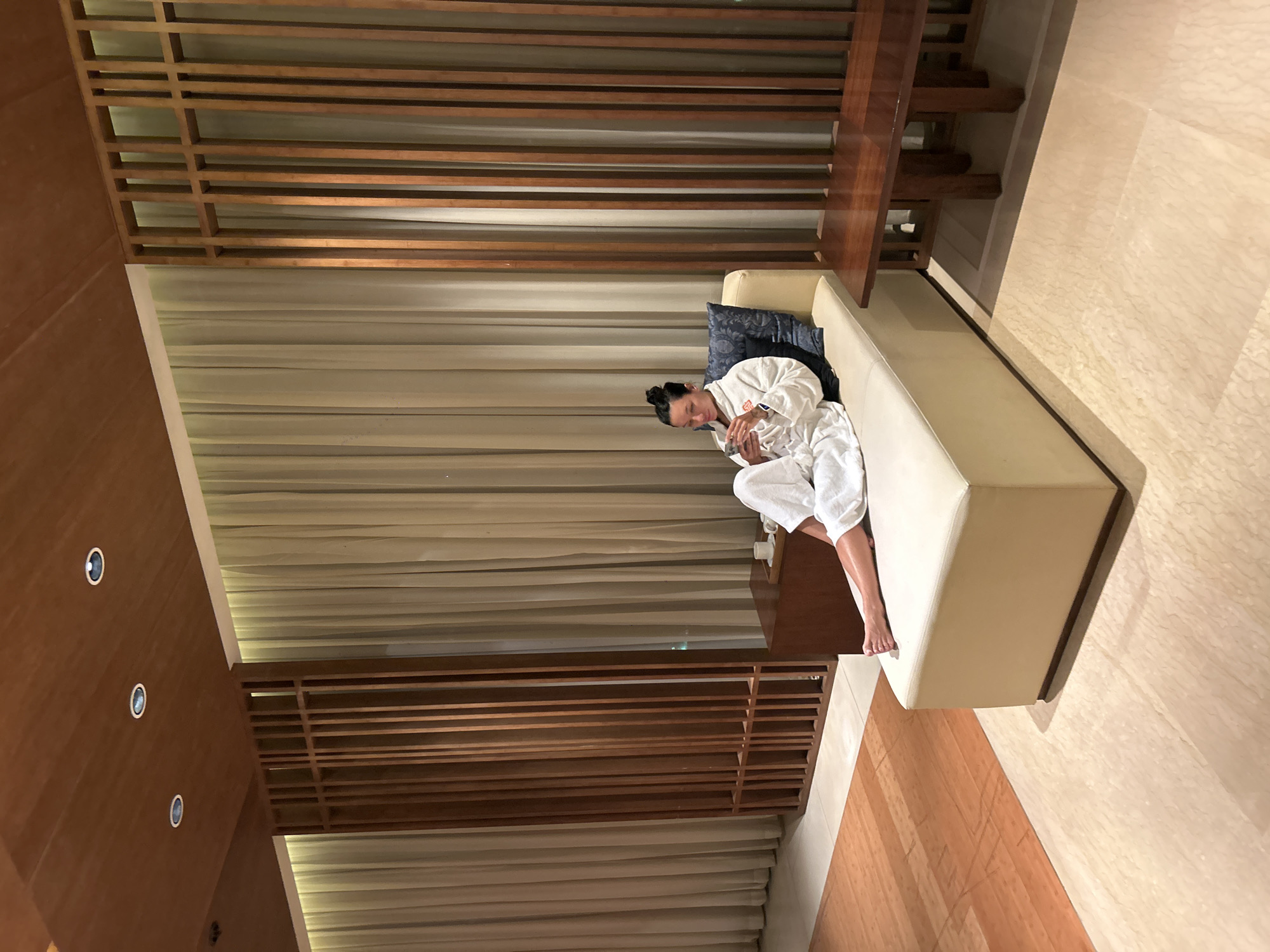 The image depicts a woman in a white bathrobe sitting on a cream-colored chaise lounge. She is holding a smartphone and appears to be looking at it. The chaise lounge is placed in a room with a wooden ceiling and wooden slatted partitions, suggesting a modern spa or relaxation area. Cream-colored curtains hang in the background. To the left of the chaise lounge, there is a small wooden table with a tray holding two cups. To the right, there is a dark wooden bench. The floor is made of light-colored patterned tiles, and an orange rug is visible on the left side of the frame. The overall impression is one of calm and relaxation. The image does not contain any elements that would allow for the determination of its location in Pune, India.