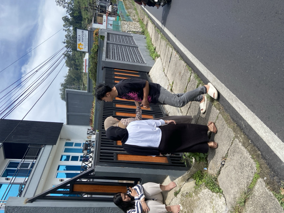 The image shows a group of four people standing in front of a gate. The people are all wearing casual clothing and appear to be engaged in conversation. Two people are wearing hijabs, one is wearing a black skirt, and one is wearing jeans. The gate appears to be a part of the property of a house, the facade of which is visible in the background. The house is painted white and has large windows. A sign reading "VILLA HAPOSAN" is visible on the side of the house. The scene is located in front of a house on a street in the city of Rancabali, Indonesia, at the coordinates -7.121 latitude and 107.433 longitude. The road is paved and has a white line down the center. There is greenery and a patch of dirt on the side of the road, and a motorcycle is visible further down the road. The sky is blue and there are clouds visible.