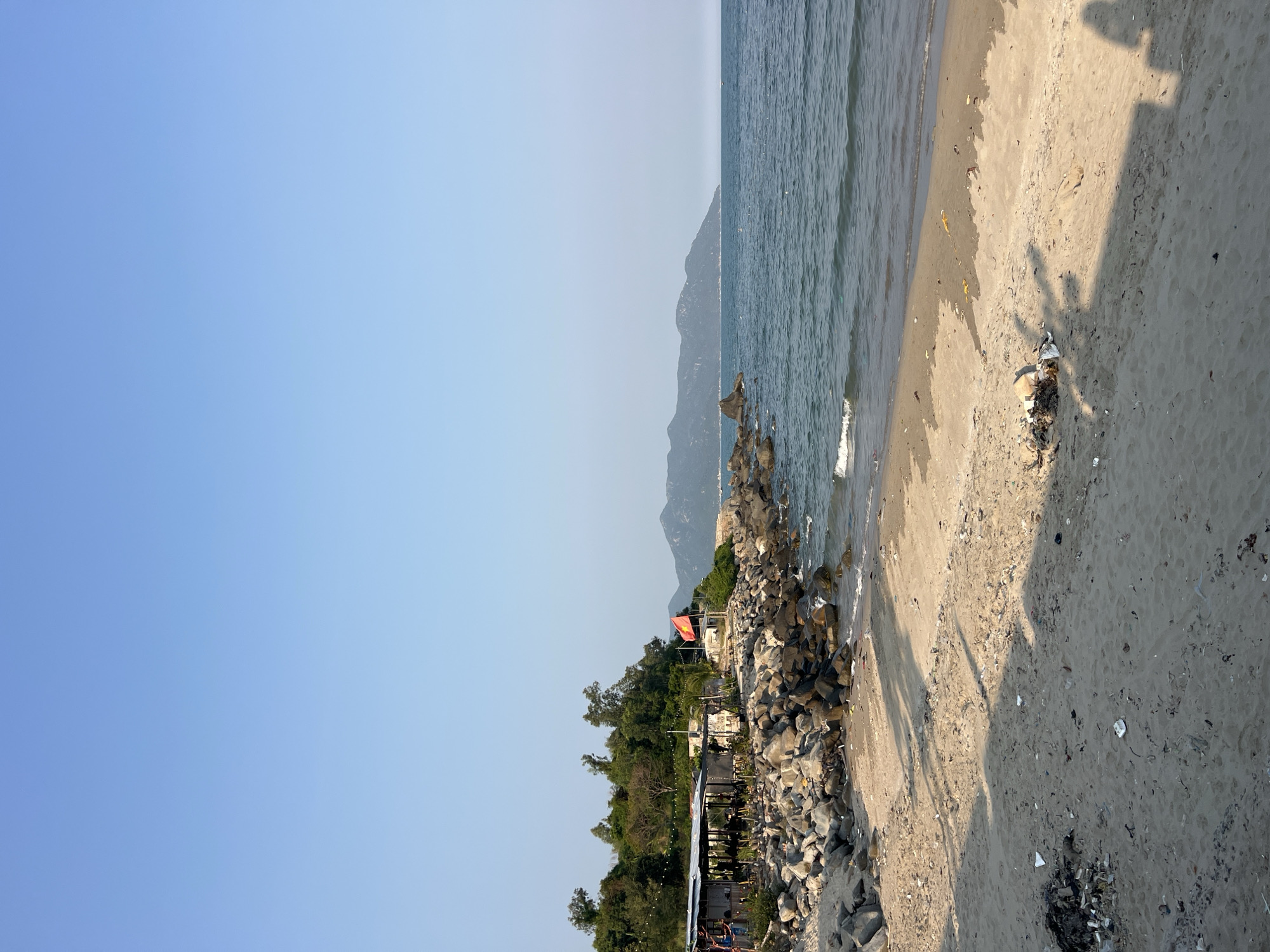 The image captures a coastal scene in Vĩnh Lương, Vietnam. A sandy beach occupies the right portion of the frame, with trash scattered across the sand. The shadow of a person is cast on the beach. To the left, the water meets the shore, with rocks lining the edge. Beyond the rocks, there's vegetation and structures including a building with an orange roof. In the distance, hills are visible, and the sky is clear blue.