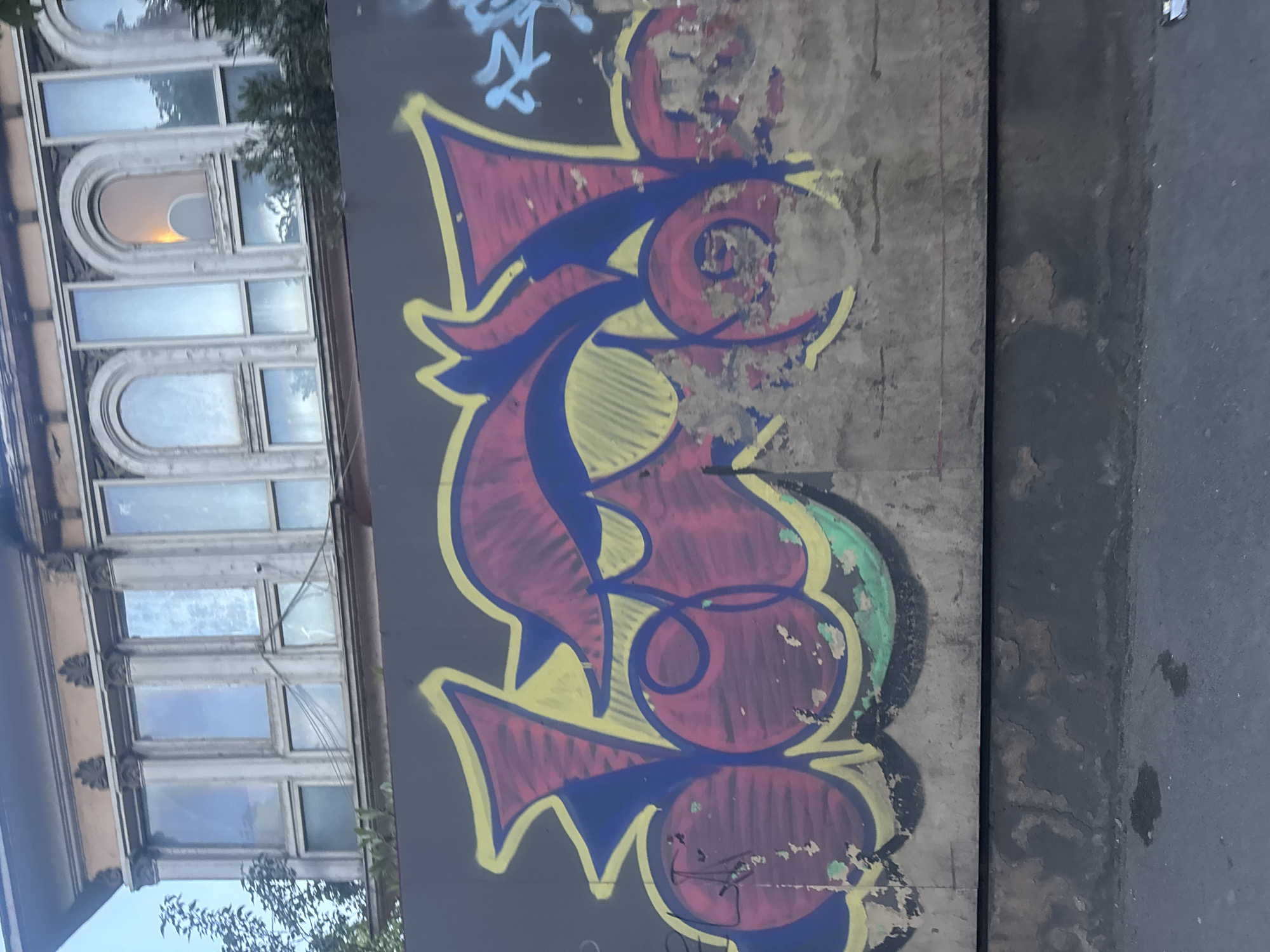 The image shows a gray wall with colorful graffiti in Tbilisi, Georgia. The graffiti is a stylized design in shades of red, yellow, and blue. The background shows a building with arched windows and some greenery. The edge of a road or street is visible on the right side.