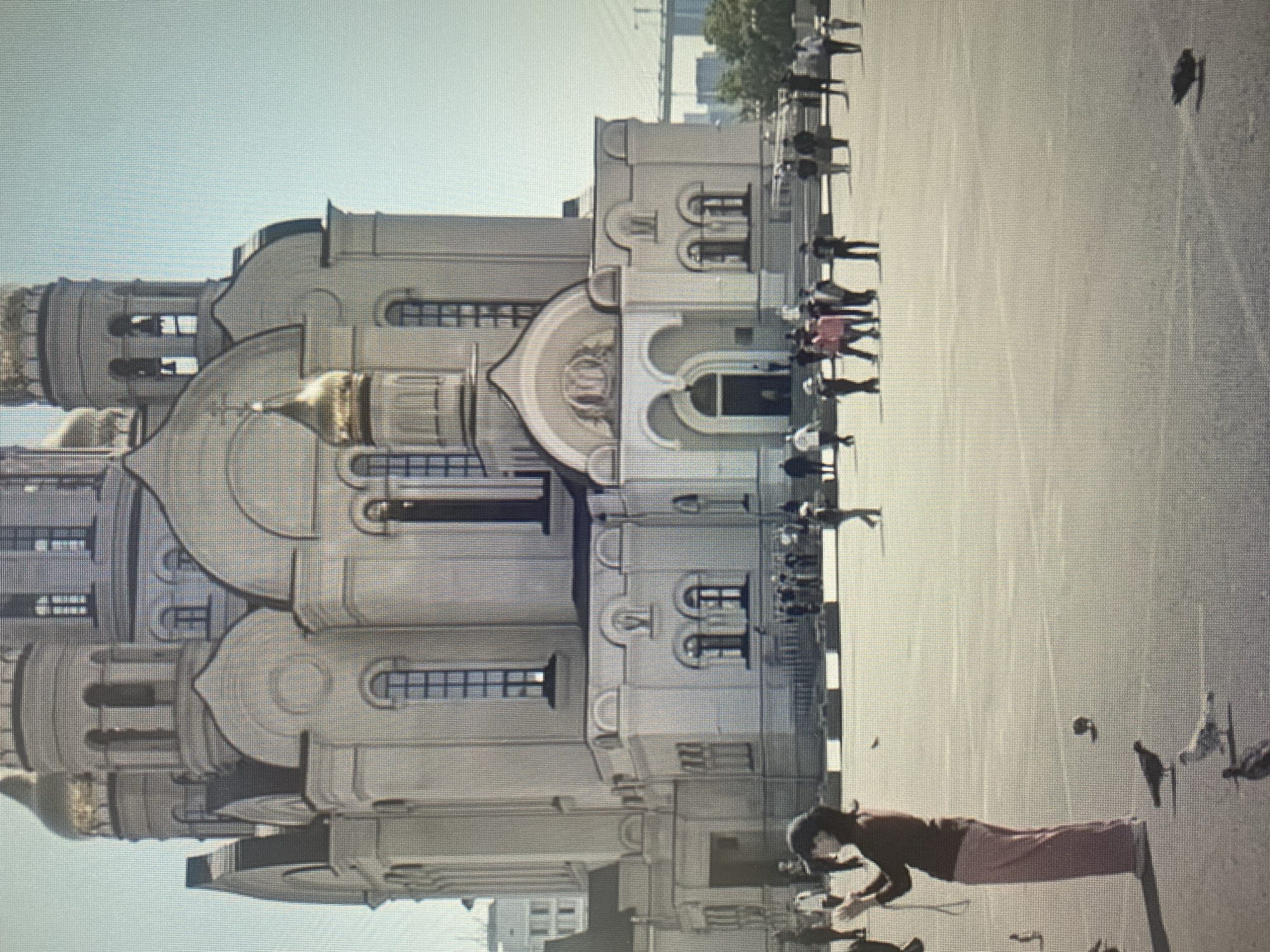 The image shows a large, ornate cathedral with multiple golden domes against a clear blue sky. The cathedral has a pale, beige facade with arched windows and intricate architectural details. In the foreground, a plaza is visible with several people walking and standing around. A woman in the foreground is holding a phone, seemingly taking a picture or recording. There are also several pigeons on the ground. In the background, a bridge and distant buildings can be seen.
This image is of the Holy Assumption Cathedral in Vitebsk, Belarus.