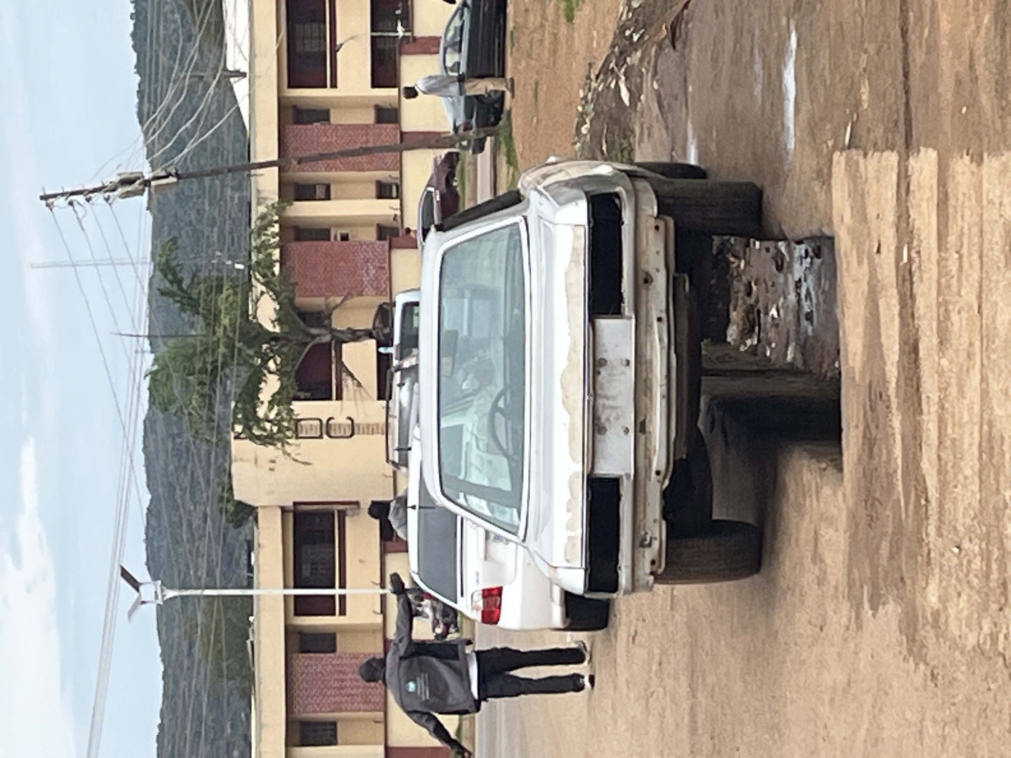 The image depicts a street scene, likely in Rafin Par, Nigeria, with a focus on several vehicles and people. In the foreground, the rear of a silver, possibly abandoned or dismantled, car is visible, positioned over a dark, open drainage ditch filled with debris. To the left, a man wearing a dark jacket with a white logo and dark pants stands with his arms outstretched, seemingly gesturing or directing. Behind him, a white car is parked, and further back, another person can be seen. The background features a long, beige building with red brick accents and numerous windows. A utility pole with multiple wires is prominent on the right side of the image, and beyond that, a green hill or mountain range is visible under a cloudy sky. Other vehicles, including a dark sedan, are present on the street. The ground is a mix of dirt and what appears to be a paved or semi-paved surface.