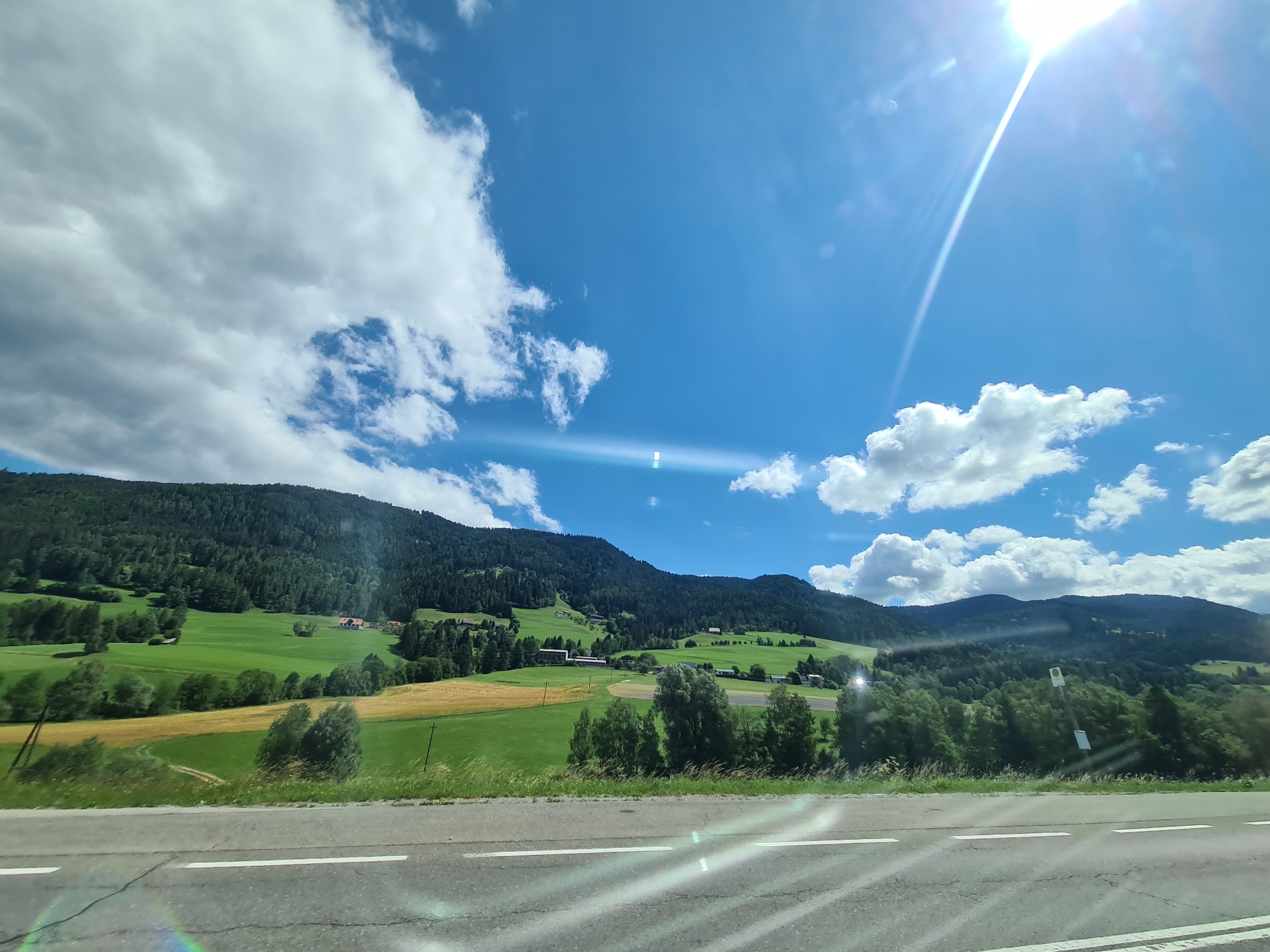 The image depicts a scenic landscape in Perchau am Sattel, Austria. In the foreground, a paved road with white lane markings runs horizontally across the frame. Beyond the road, a lush green meadow slopes upwards towards a forested mountain range. Several farmhouses with red roofs are nestled on the hillsides. The sky is a vibrant blue, dotted with white, fluffy clouds. The sun is shining brightly, casting lens flares across the image. The overall impression is one of a serene, rural setting bathed in natural light.
