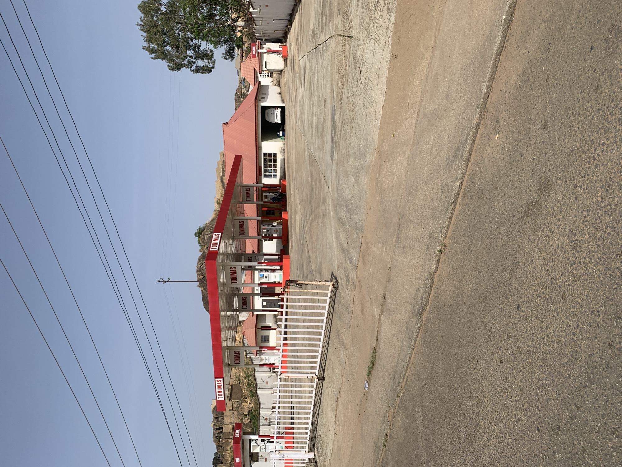 The image shows a TOMIMAS gas station in Jos, Nigeria. The station has a red and white color scheme. There are visible gas pumps under a red awning. A white gate is positioned near the pumps, and electrical wires are visible in the sky. The terrain surrounding the gas station is rocky and hilly. The sky is clear and light blue. A concrete road and parking area are adjacent to the station. There is a one story house adjacent to the gas station with a red roof. An auto repair shop is visible, and a white car is in the shop.