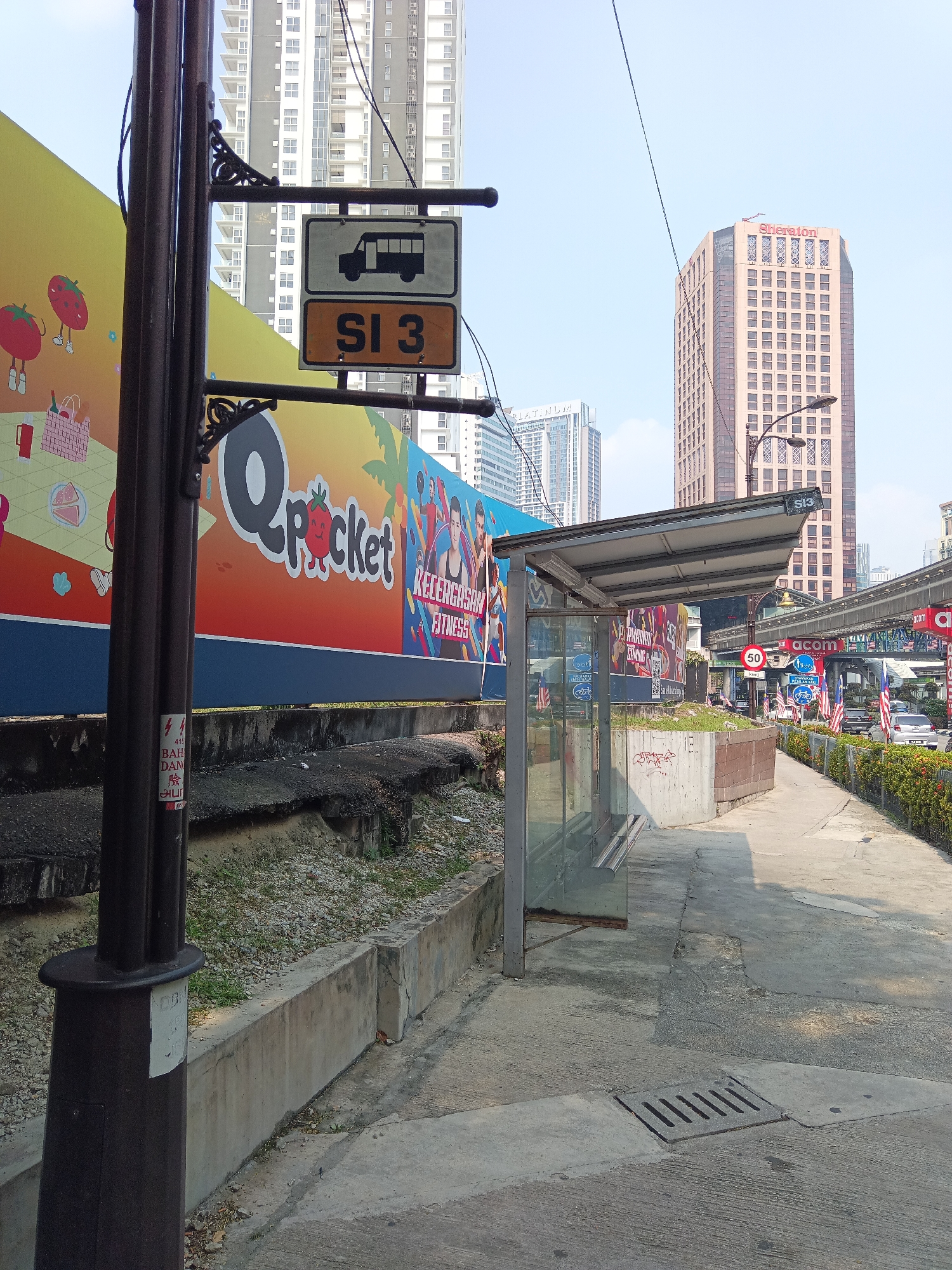 The image depicts a bus stop scene in Kuala Lumpur, Malaysia, with a prominent bus stop sign displaying "SI 3". A bus stop shelter with glass panels and a metal roof is visible on the right. Behind the shelter, a large, colorful advertisement featuring cartoon strawberries and the text "Qpocket" and "RECREATION FITNESS" is displayed. Several tall buildings, including the Sheraton hotel, are seen in the background under a clear blue sky. A monorail track runs overhead, with signs indicating "acm". The street is paved, with a drain grate in the foreground. The general environment suggests an urban setting with ongoing commercial activity.
