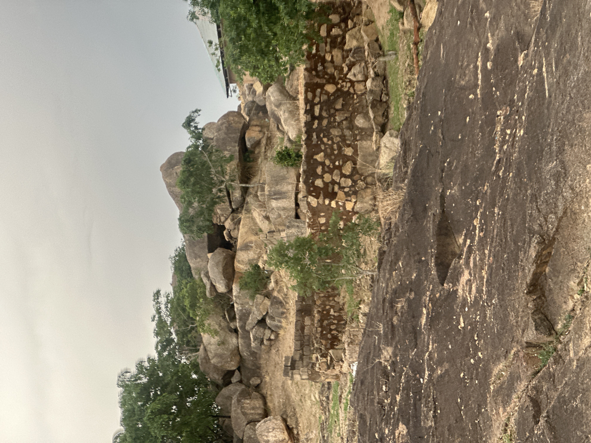 The image shows a rocky landscape, potentially a hillside or cliff, under an overcast sky. The scene features large boulders and rock formations, with some green vegetation, including trees and shrubs, interspersed among the rocks. A section of a rock wall is visible. The photo might depict a location around Jos, Nigeria, characterized by similar rocky terrain and vegetation. No people or animals are visible.