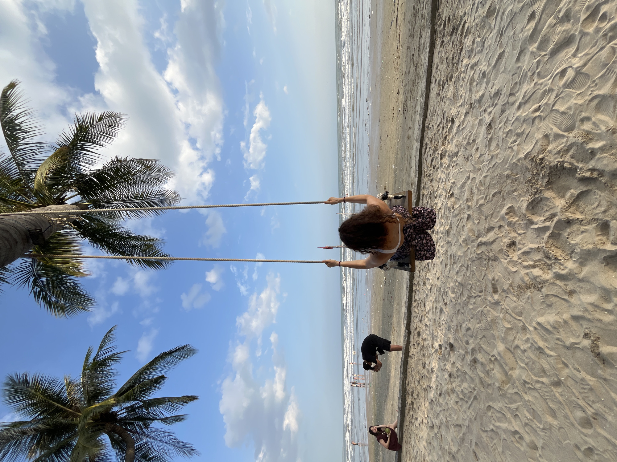 The image depicts a beach scene in Mũi Né, Vietnam, where a woman is seated on a swing made of rope and wood, facing the ocean. She has long, curly brown hair and is wearing a patterned skirt and a white top. The swing is suspended from a palm tree, whose fronds are visible in the upper part of the frame against a blue sky with scattered white clouds.
In the background, the ocean stretches to the horizon, with waves breaking on the shore. The beach is sandy, with footprints visible in the foreground. Several other people are present on the beach: a woman in a dark dress is sitting on a low wall in the mid-ground, holding a coconut; a man in a black t-shirt and shorts is bending down, appearing to take a photo with his phone; and other figures can be seen further down the beach near the water. The overall atmosphere is relaxed and indicative of a beachside resort or public beach.