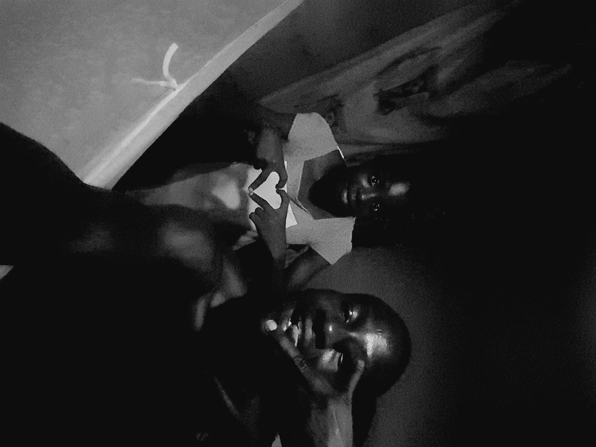 The image is a low-light, black and white photograph featuring two individuals. In the foreground, a person is partially visible, their face illuminated and looking towards the camera. They are making a gesture with their hand over their eye, possibly pointing or posing. Behind them and slightly to the left, a second individual is also visible, looking directly at the camera with wide eyes. This person is forming a heart shape with their hands. The background is dark, with some indistinct shapes that might suggest an indoor environment. The image appears to be taken in Narayi, Nigeria, though no specific landmarks or environmental details confirm this location. The overall atmosphere is intimate and informal, characteristic of a personal photograph.
