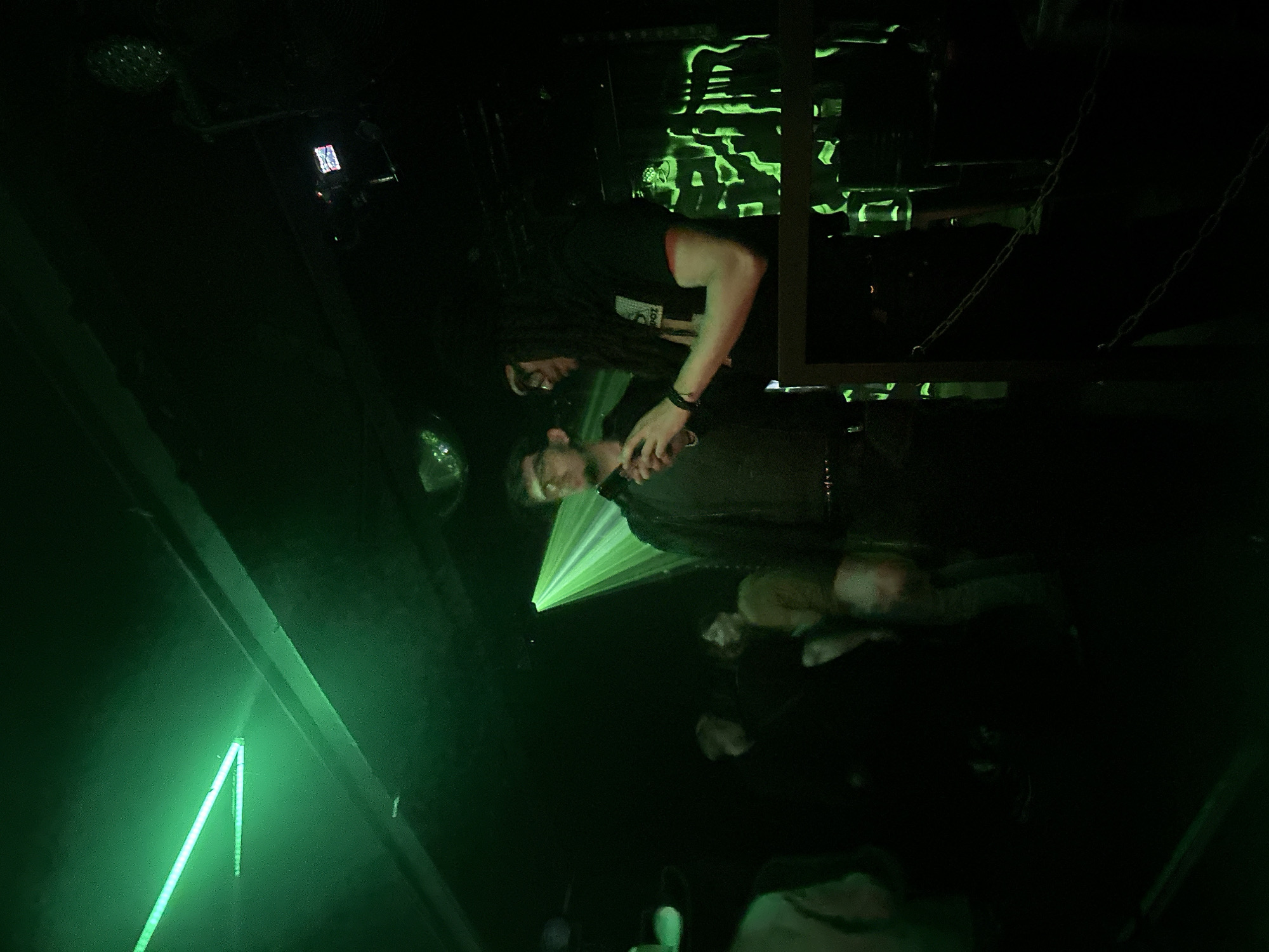 The image depicts an indoor scene within a dimly lit nightclub or music venue. Green light dominates the environment, originating from linear LED strips on the ceiling, a conical spotlight illuminating the central figures, and projected patterns on a background wall. A partially visible disco ball hangs from the ceiling. In the mid-ground, two men stand behind a dark railing featuring two draped chains. The man on the left wears glasses, a dark jacket, and holds an object resembling a smartphone. He looks towards the right. The man on the right has long dark dreadlocks or braids, wears a dark t-shirt, and is actively manipulating a smartphone with both hands. Multiple other individuals are present in the blurred background, some partially visible, including a woman in a light brown top and silhouetted figures against the green wall projections. The specific location within the city of Paris, France, cannot be determined from the visual information provided.