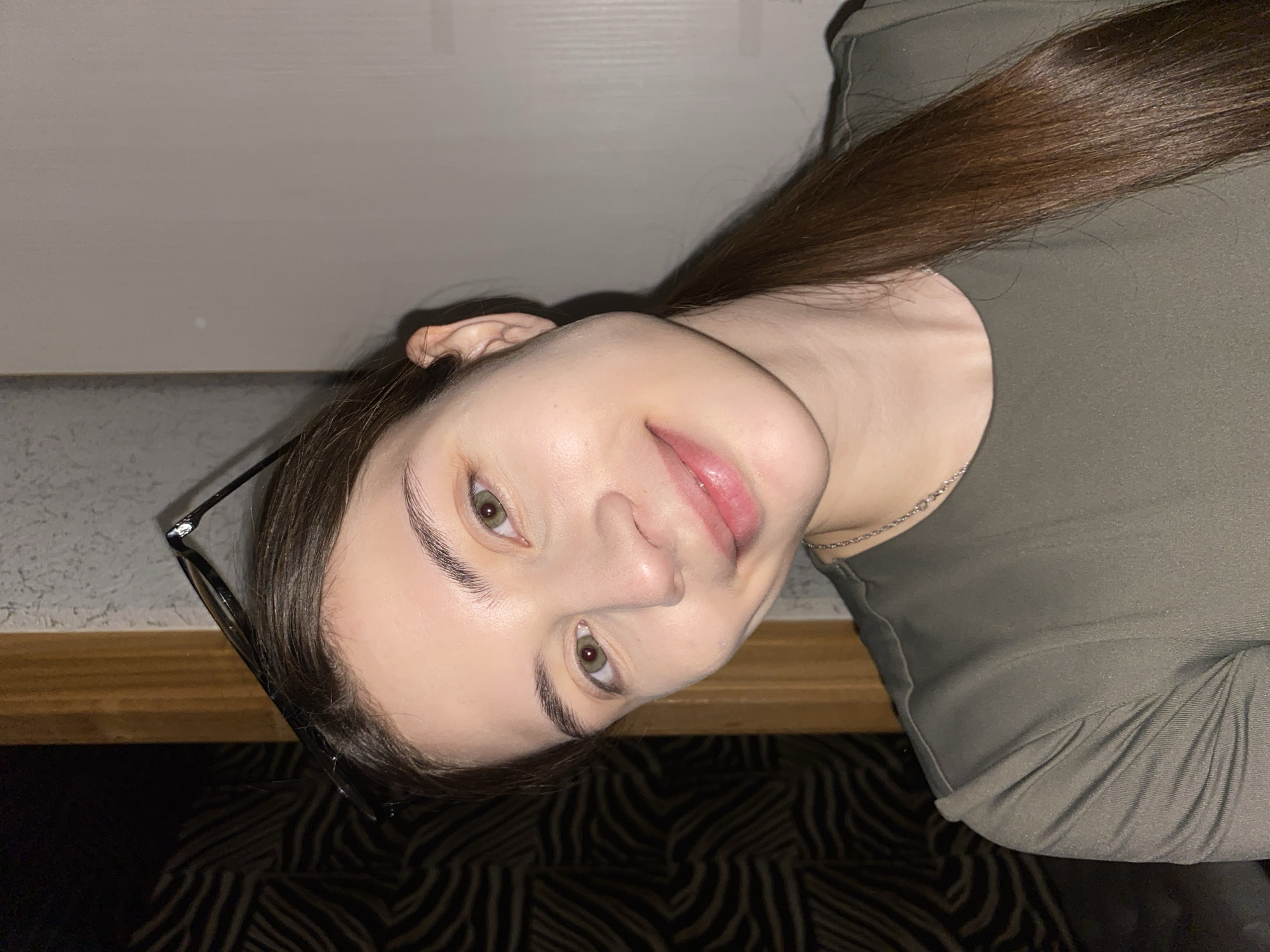 This media file captures a close-up, rotated portrait of a young woman taken indoors in Votkinsk, Russia. The image is rotated 90 degrees counter-clockwise.
**Subjects:** The primary subject is a young woman. Her face is visible, featuring clear skin, green eyes, and neatly shaped dark eyebrows. She has long, straight, dark brown hair that flows down her back and partially frames her face. Black-framed glasses are resting on top of her head or pushed up into her hair. She is wearing a fitted, dark olive-green top and a thin silver chain necklace around her neck.
**Activity or event taking place:** The woman is posing for a photo, likely a selfie, given the angle and proximity.
**Notable details:**
* **Emotions:** She has a calm expression with a slight, gentle smile.
* **Lighting:** The lighting suggests an indoor setting, possibly with artificial light, as there are no strong shadows indicative of direct natural light.
* **Background:** The background behind her head is a light, plain wall or door. Below her, the background transitions to a dark surface with a distinct, light-colored zebra-stripe or wave-like pattern, which could be upholstery, a rug, or a textured floor. A strip of wood-grained material is also visible below the light wall/door.
* **Absence of visible text:** There is no discernible text within the image.
**Scene and Setting:** The setting is an indoor space, possibly a home or private room, characterized by the simple wall and patterned background elements.