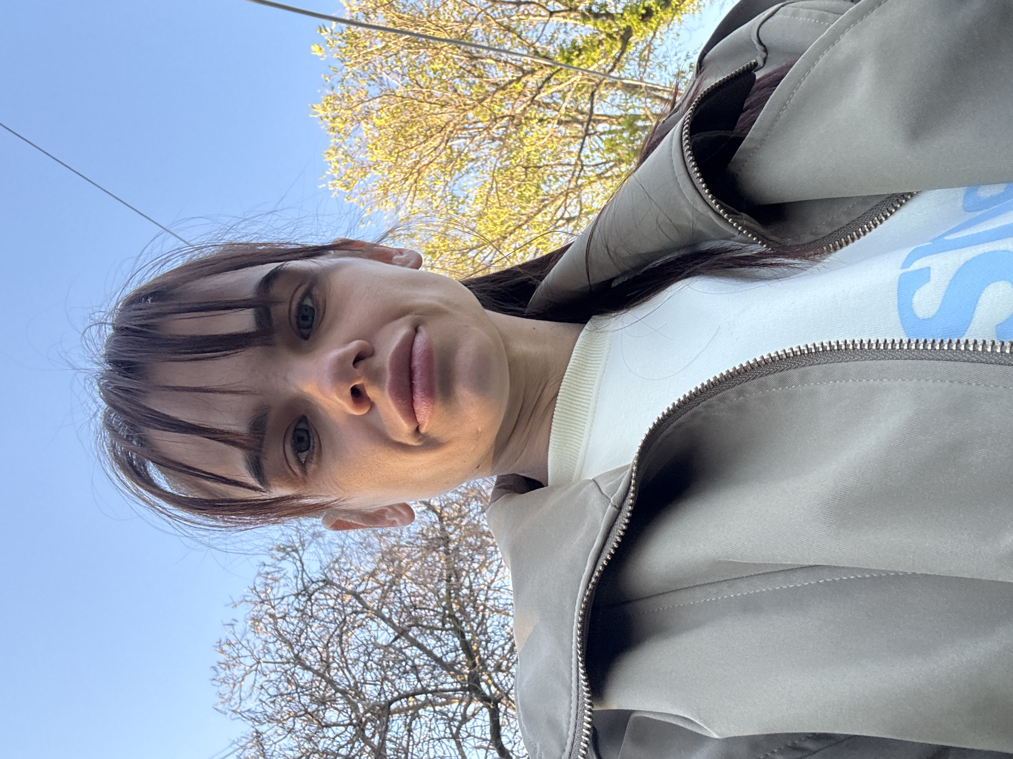 A woman with dark hair and bangs, wearing a light beige jacket and a white shirt, is looking directly at the camera. The photo is taken from a low angle, looking upwards. Above her, the sky is visible, partially obscured by the branches of trees. Although a location is requested, the image does not provide any specific features or context to determine that the location is Rostov-on-Don, Russia.
