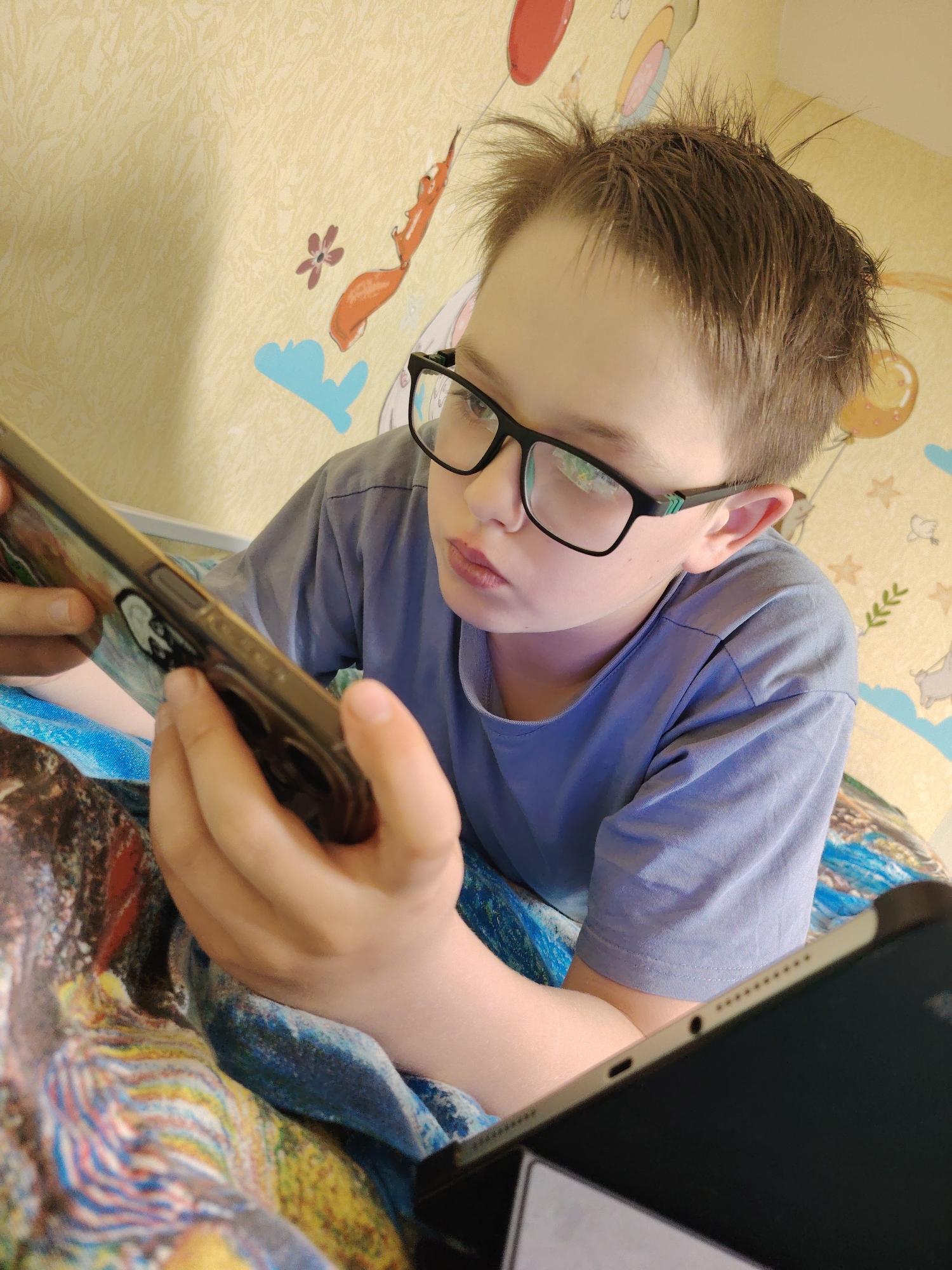 A young boy wearing black-rimmed glasses is lying down while looking at a smartphone in his hands. He is also positioned near a tablet. The background reveals a wall decorated with cartoon figures and balloons. This image was captured in a location within the city of Chaikovsky, Russia.