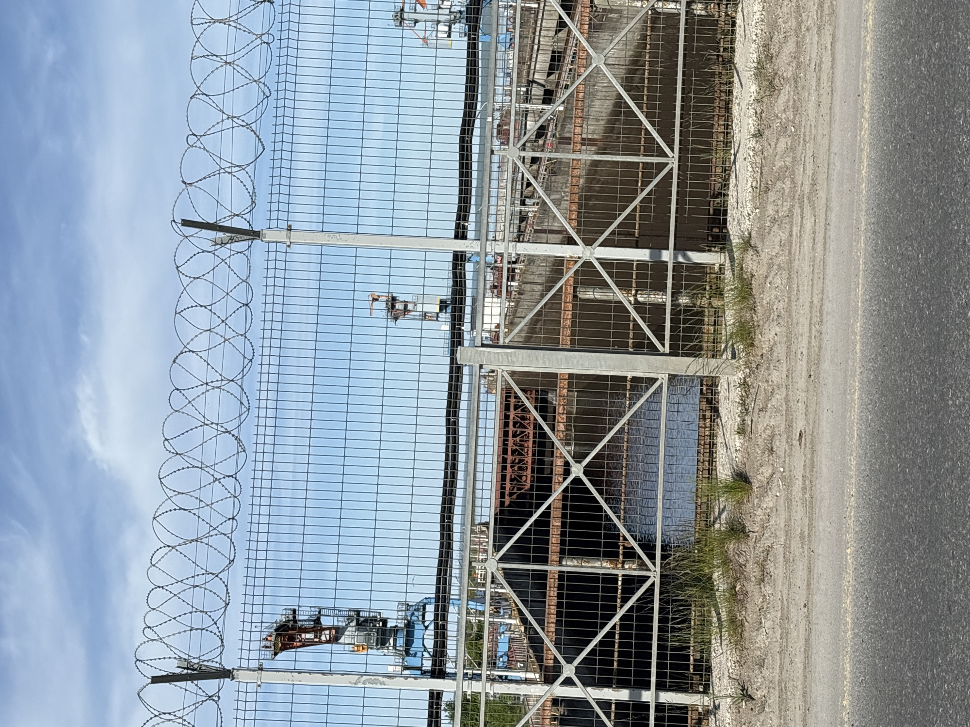 The image depicts a high-security perimeter fence with barbed wire at the top. Beyond the fence, a large industrial structure is visible through a secondary metal fence. The ground adjacent to the fence appears to be a mix of dirt, gravel, and asphalt. The sky is partially visible behind the fence, suggesting an outdoor location. Based on the city mentioned, the scene is located near a significant industrial complex, likely a factory or energy production site in Chaikovsky, Russia. No people are visible.