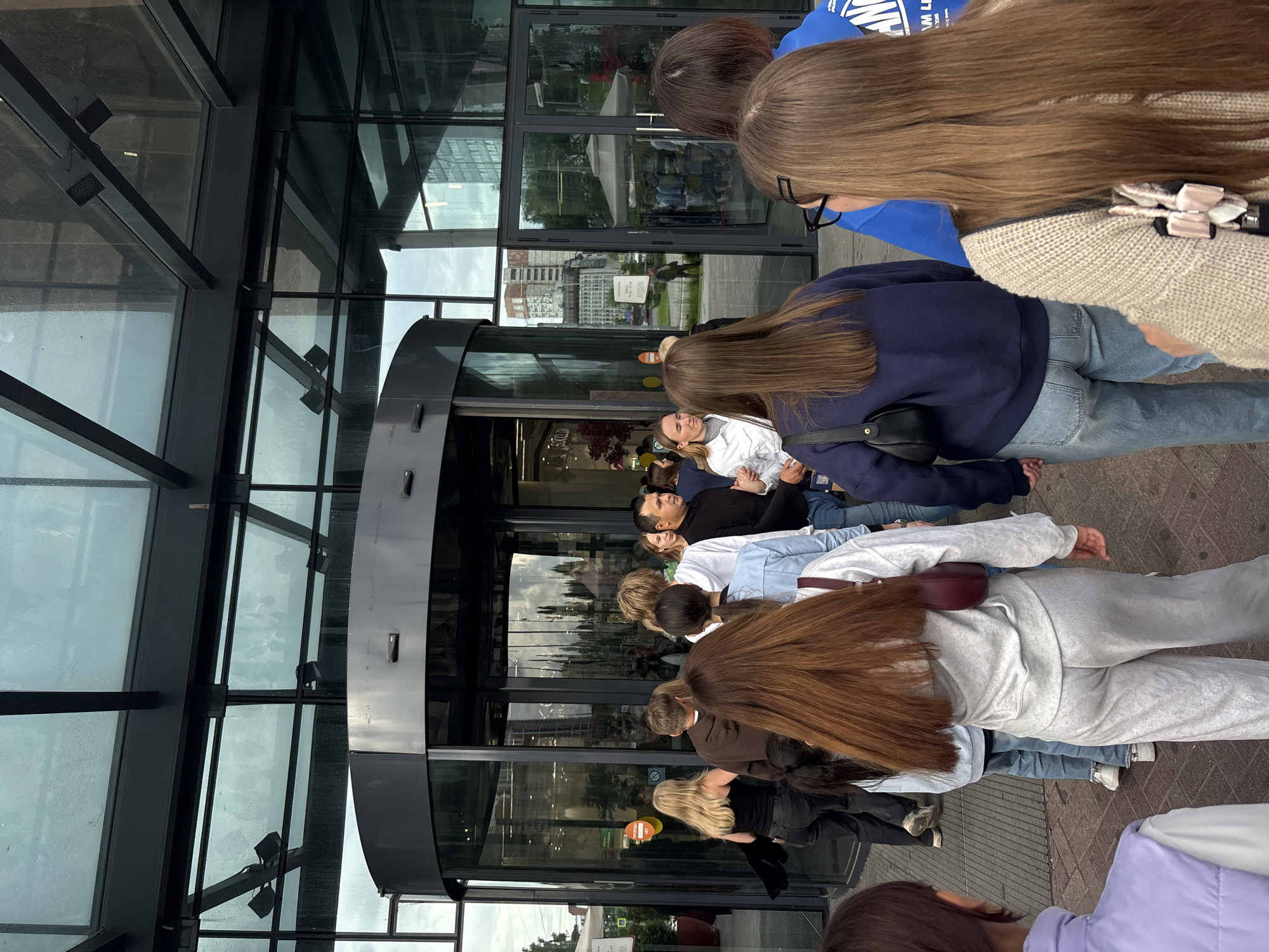 A queue of approximately 15-20 individuals, predominantly young adults, is depicted at the entrance of a modern glass-fronted building in Perm, Russia, under an overcast sky. The primary entry point is a large, dark-framed glass revolving door positioned centrally. To the right of the revolving door are two standard glass swing doors. An extensive glass canopy with dark metal framing and integrated spotlights extends over the entrance.
On the left side of the revolving door, a red-orange circular sticker with white Cyrillic text, "ПРОХОДА НЕТ" (No Passage), is visible. The ground immediately in front of the entrance is paved with interlocking concrete pavers.
In the foreground, several individuals are observed from behind. One person with long reddish-brown hair wears a light grey long-sleeved top and matching wide-leg pants, carrying a dark red shoulder bag. To their left, another individual with shoulder-length brown hair wears a dark blue long-sleeved top and light blue jeans, holding a black shoulder bag. Further back, a blonde female in a black top approaches the revolving door. To the right of the central queue, a male with short dark hair in a black long-sleeved shirt is present, along with a blonde female in a white long-sleeved top.
Reflections in the building's glass facade reveal an urban environment, including another building that appears to be under construction or renovation, and a large grey outdoor umbrella structure to the right. A partially visible sign reflecting in the glass reads "S.K.O.L.O.V.", indicating a commercial establishment within or nearby the building.