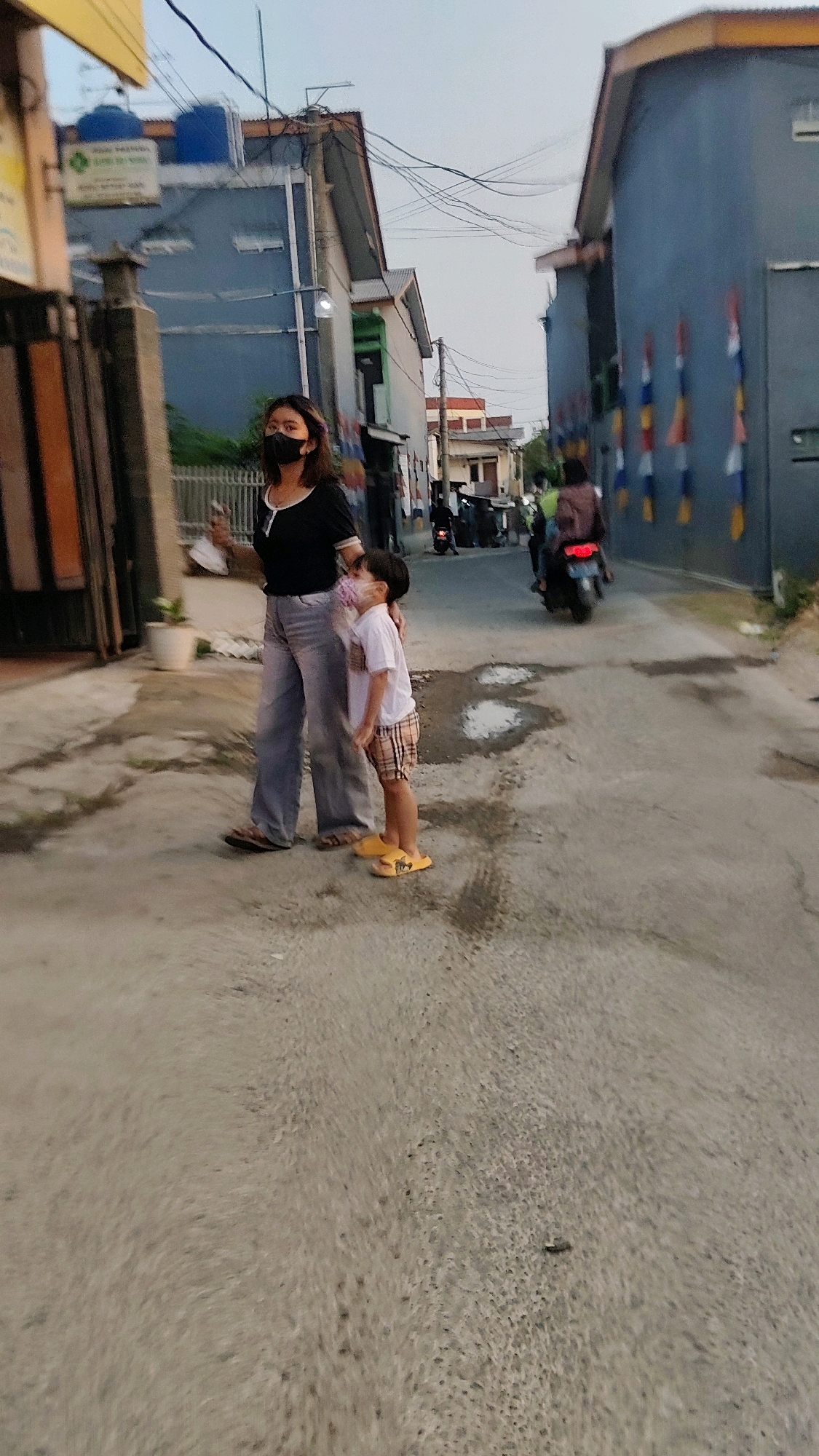 The image depicts a street scene in Mekarmukti, Indonesia, at dusk. A woman wearing a black t-shirt, light-colored pants, and a black face mask stands with a young child who is wearing a white t-shirt and plaid shorts. The child also appears to be wearing a face mask. They are standing on a wet, paved road, with puddles reflecting the sky. In the background, there are buildings with blue and grey facades, some adorned with colorful flags. A person on a motorcycle is visible further down the street, and there are power lines overhead. The overall impression is that of a residential street in a town.